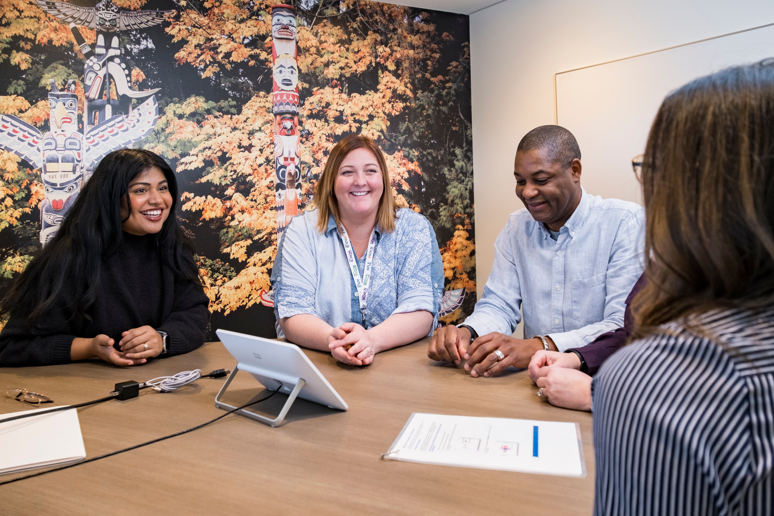 Three women and one man are meeting together at an office desk with a tablet in front of them. They are all smiling. 