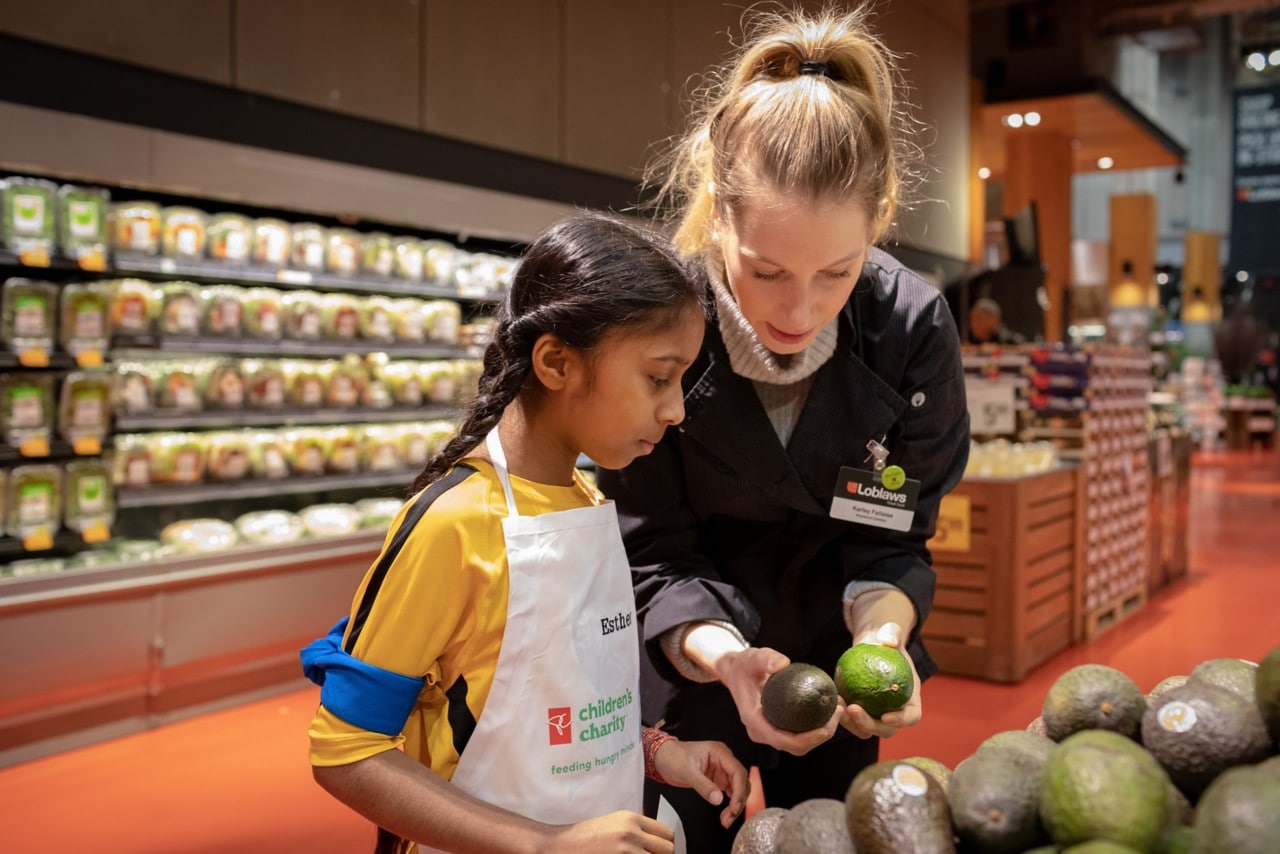 A Loblaws employee is picking out avocados with a little girl in a grocery store. 