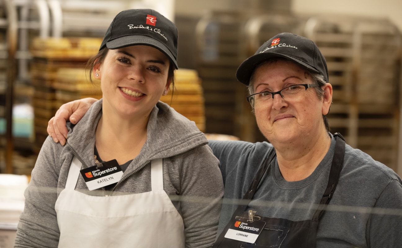 An older grocery store employee with her arm around a younger grocery employee. Both of them are smiling.