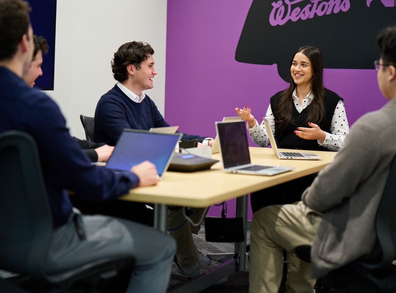 Group of interns sitting at a table with laptops listening to someone speak.