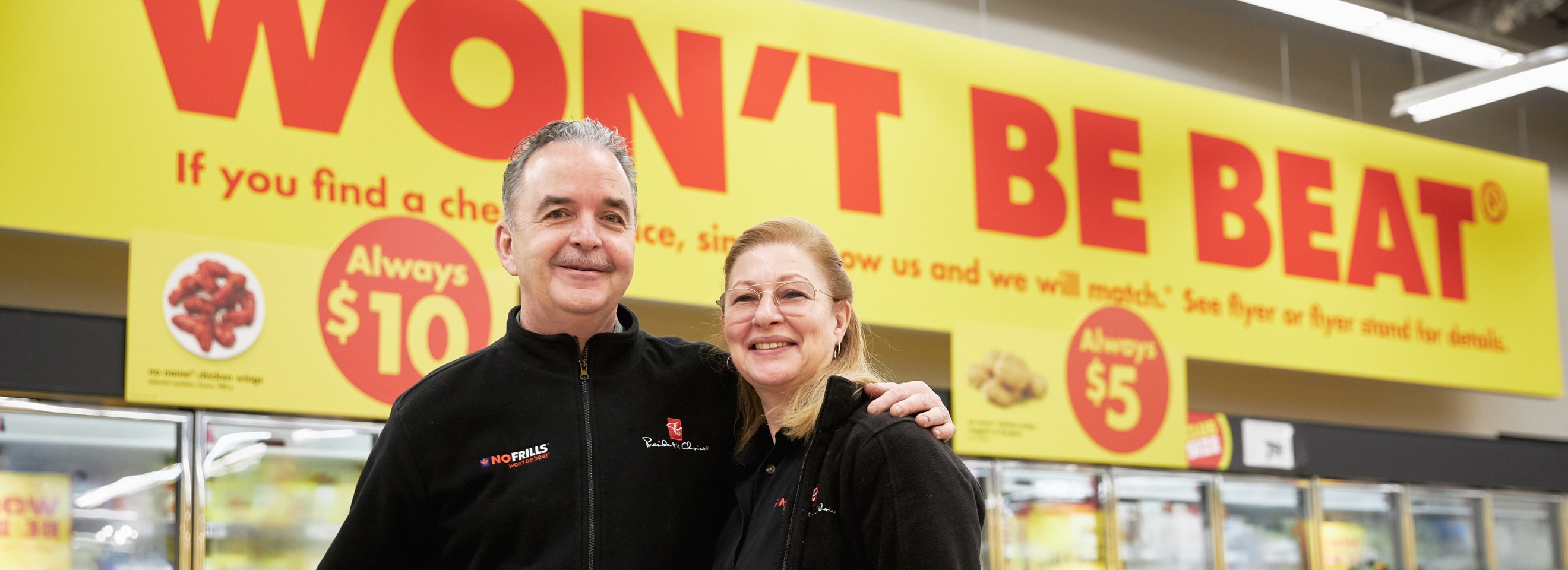 Two grocery store employees standing next to each other smiling in front of refrigerators.