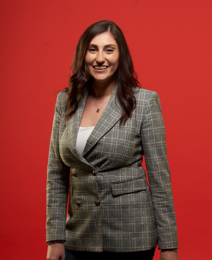 Femme portant un blazer à carreaux et souriant sur un fond rouge.