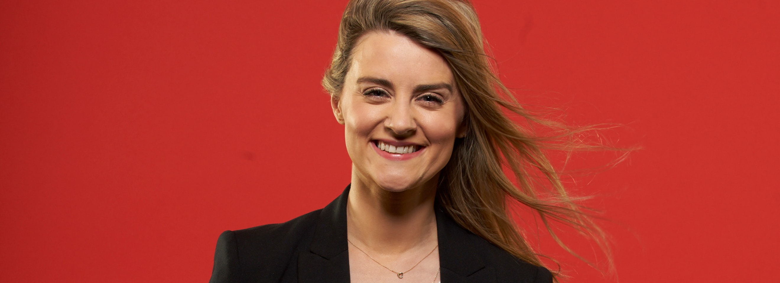 Femme portant un blazer noir et souriant sur un fond rouge.