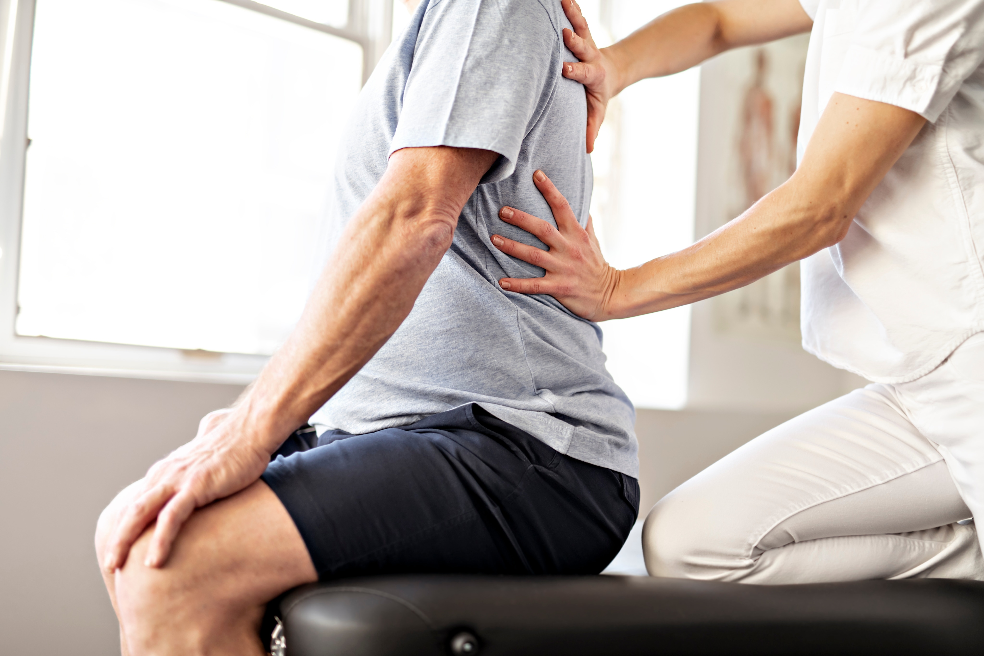 Un physiothérapeute en uniforme blanc appuie sur le dos d’un patient. Le patient est assis et le physiothérapeute est agenouillé sur le siège. 