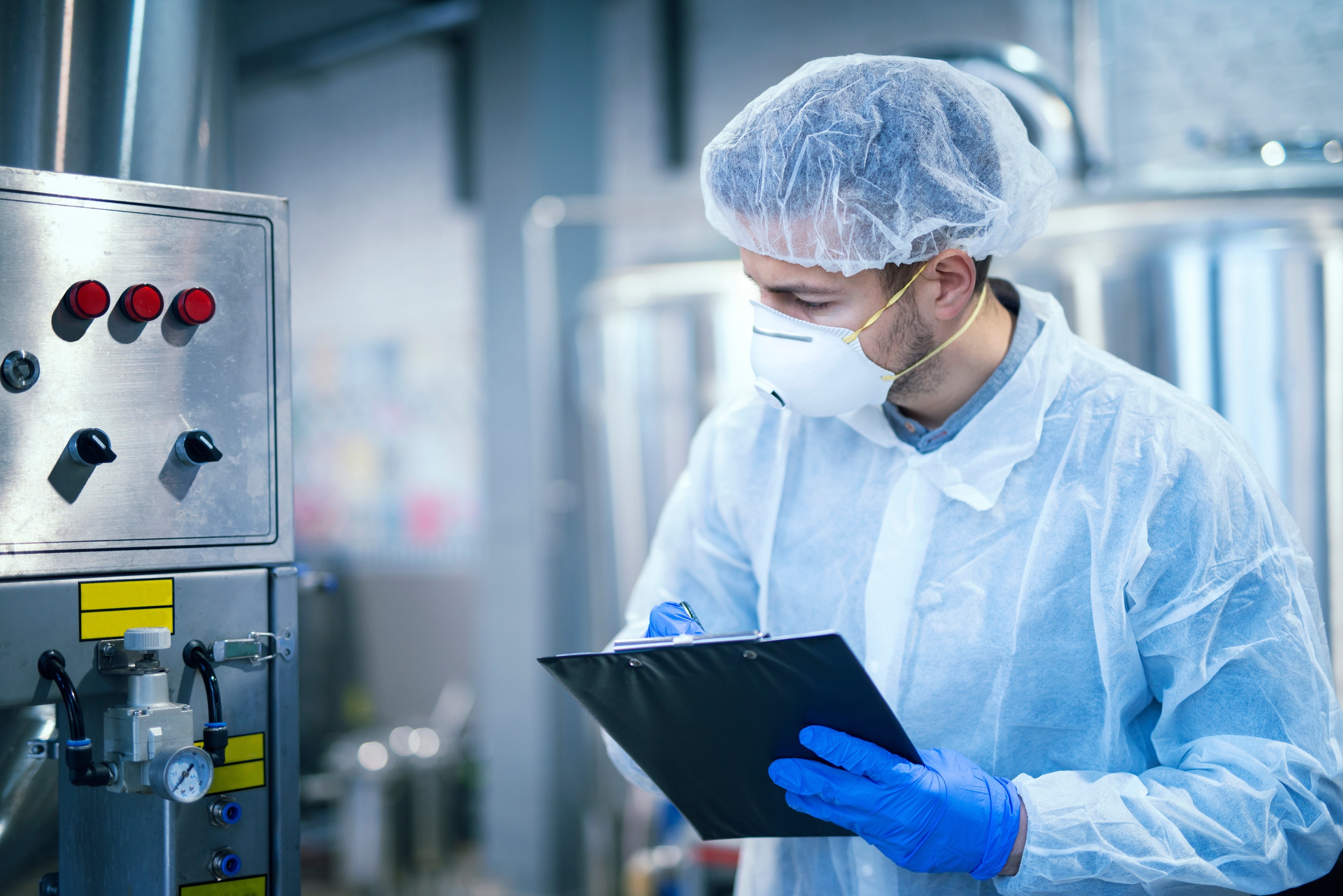 A man is taking notes with a clipboard and pen in a test lab beside a machine. He is wearing protective gear, including a hair cap, mask, shirt coverup, and gloves.