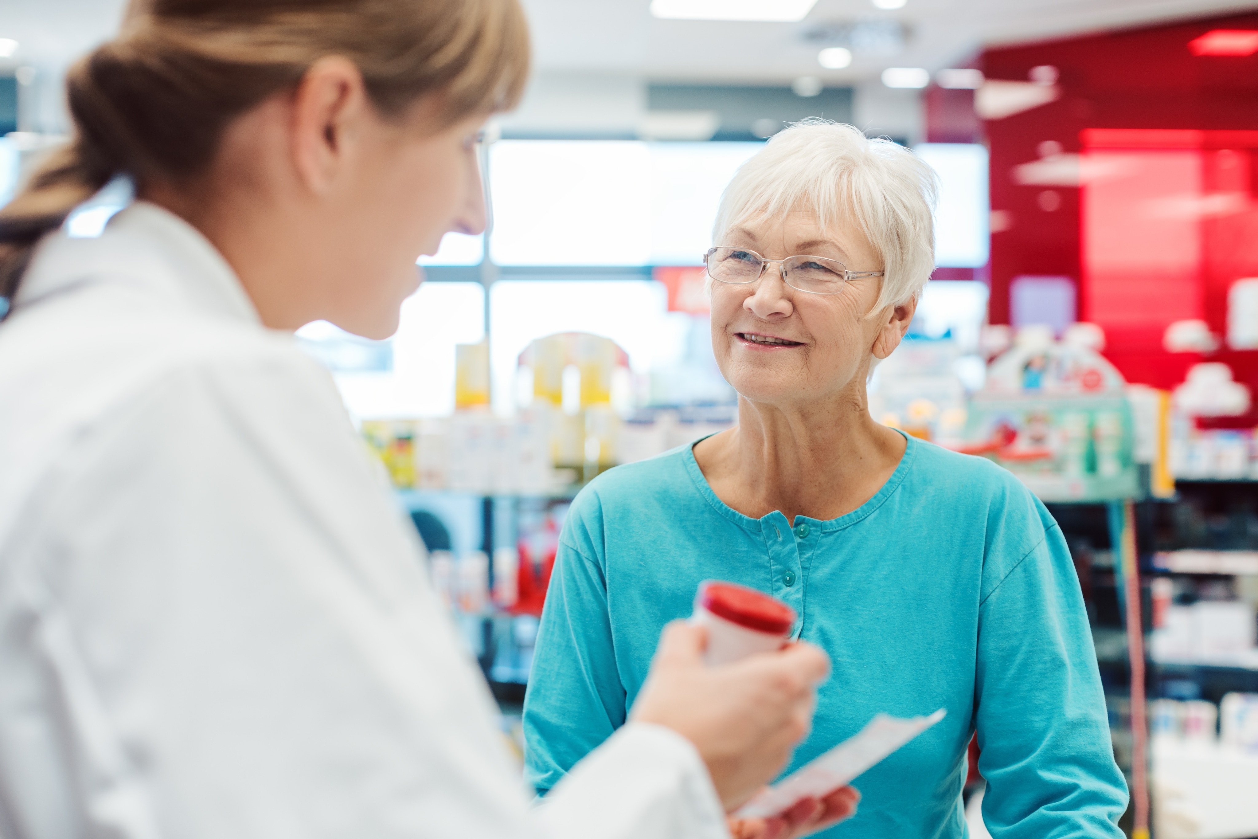 Dans une pharmacie, une pharmacienne tend un flacon de comprimés à une femme âgée. La femme âgée est attentive et souriante. 