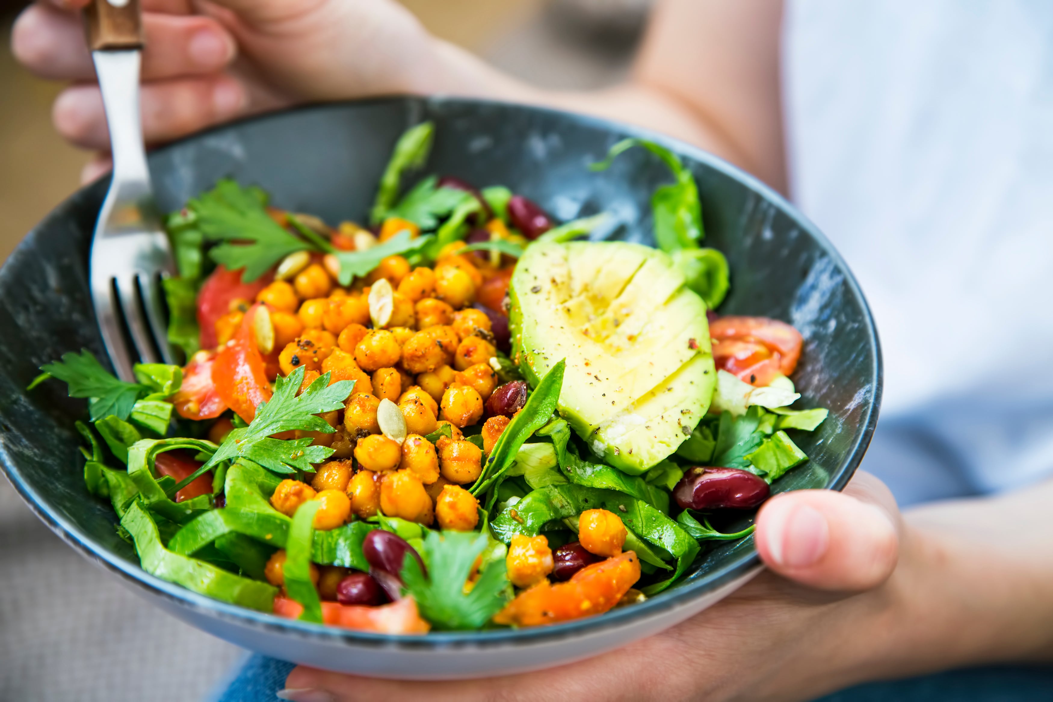 A person is holding a fork and a bowl of salad, including arugula, chickpeas, kidney beans, and avocado. 