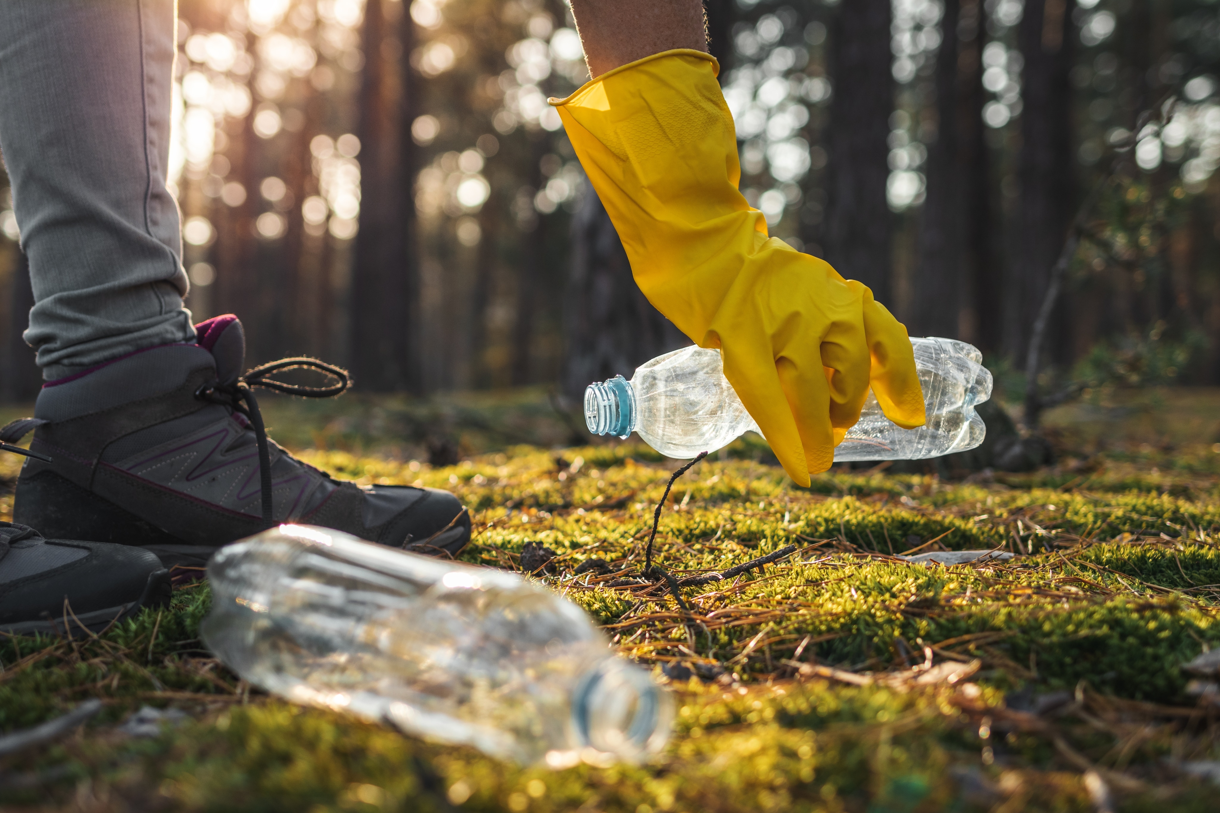 A person wearing hiking boots and a yellow glove picks up a plastic water bottle in a forest. There is another plastic water bottle nearby. 