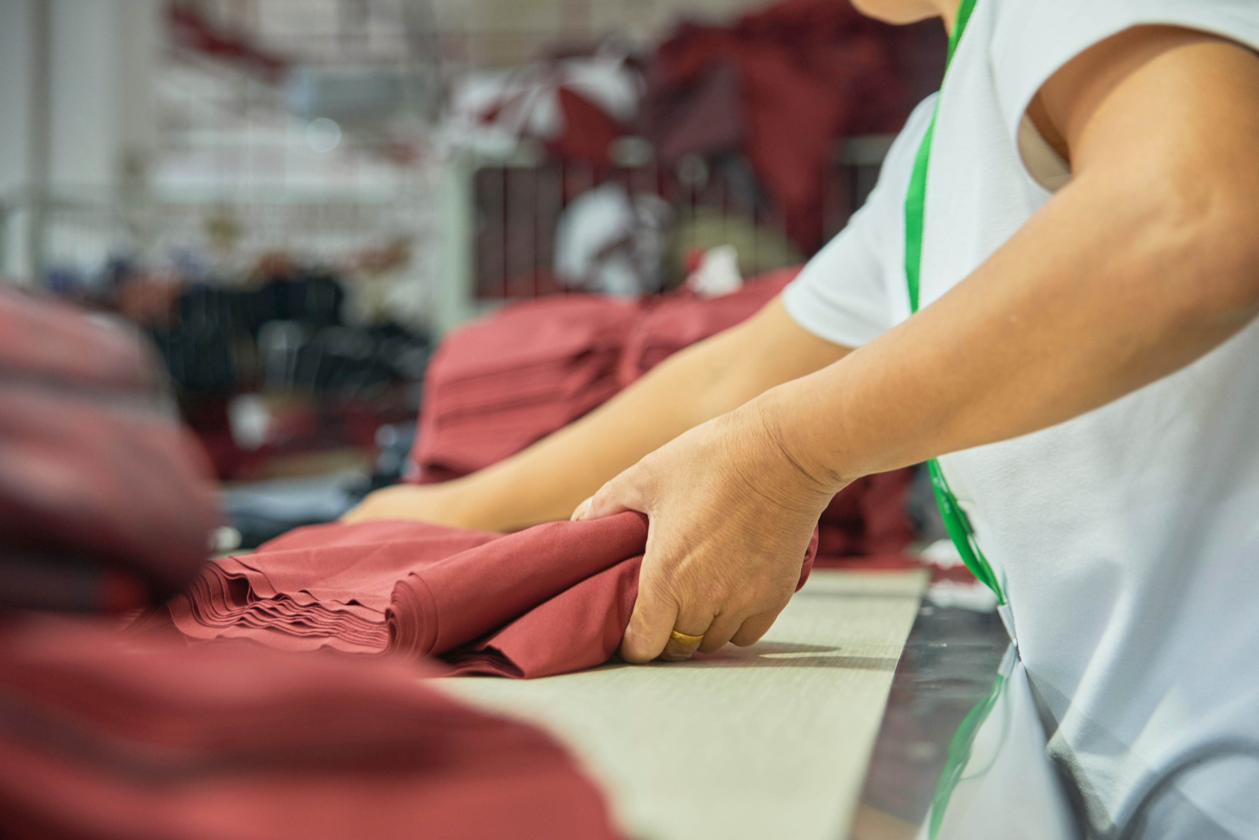 A worker in a garment factor is placing red folded fabric on a table. There are more piles of red fabric nearby.