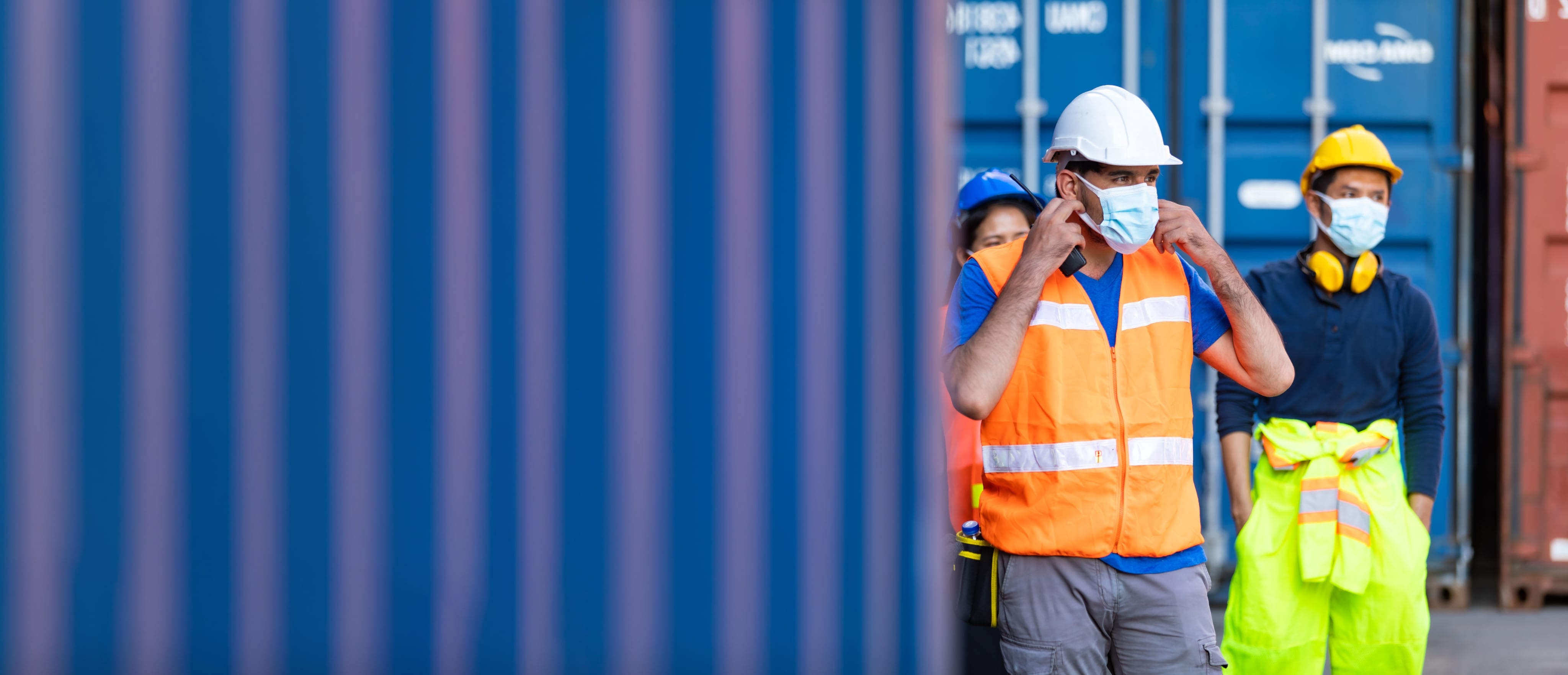 Two male workers and one female worker are wearing masks and hard hats. They are surrounded by shipping containers outside.