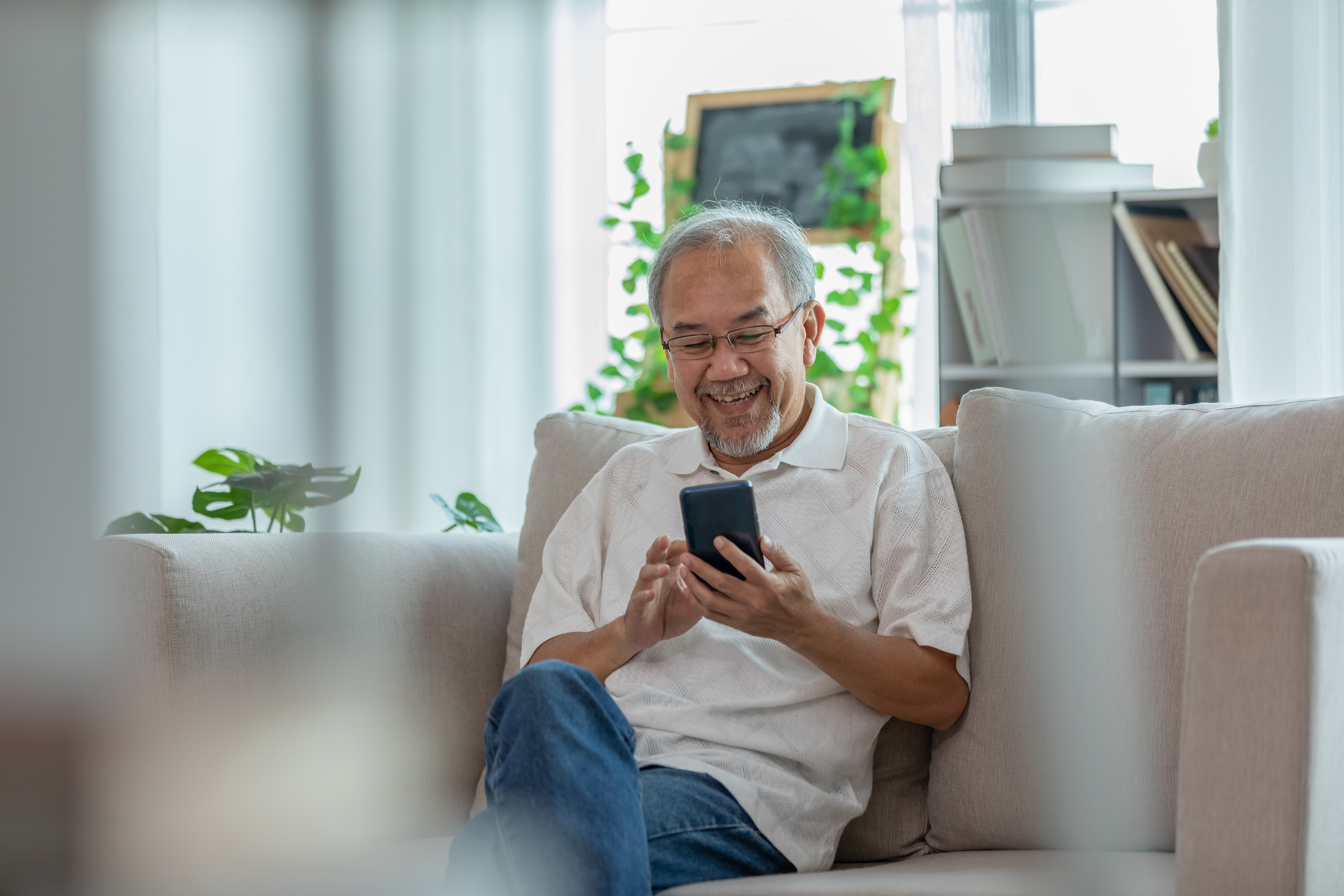 Un homme asiatique âgé est assis sur un canapé gris dans un salon. Il sourit en faisant défiler son téléphone.