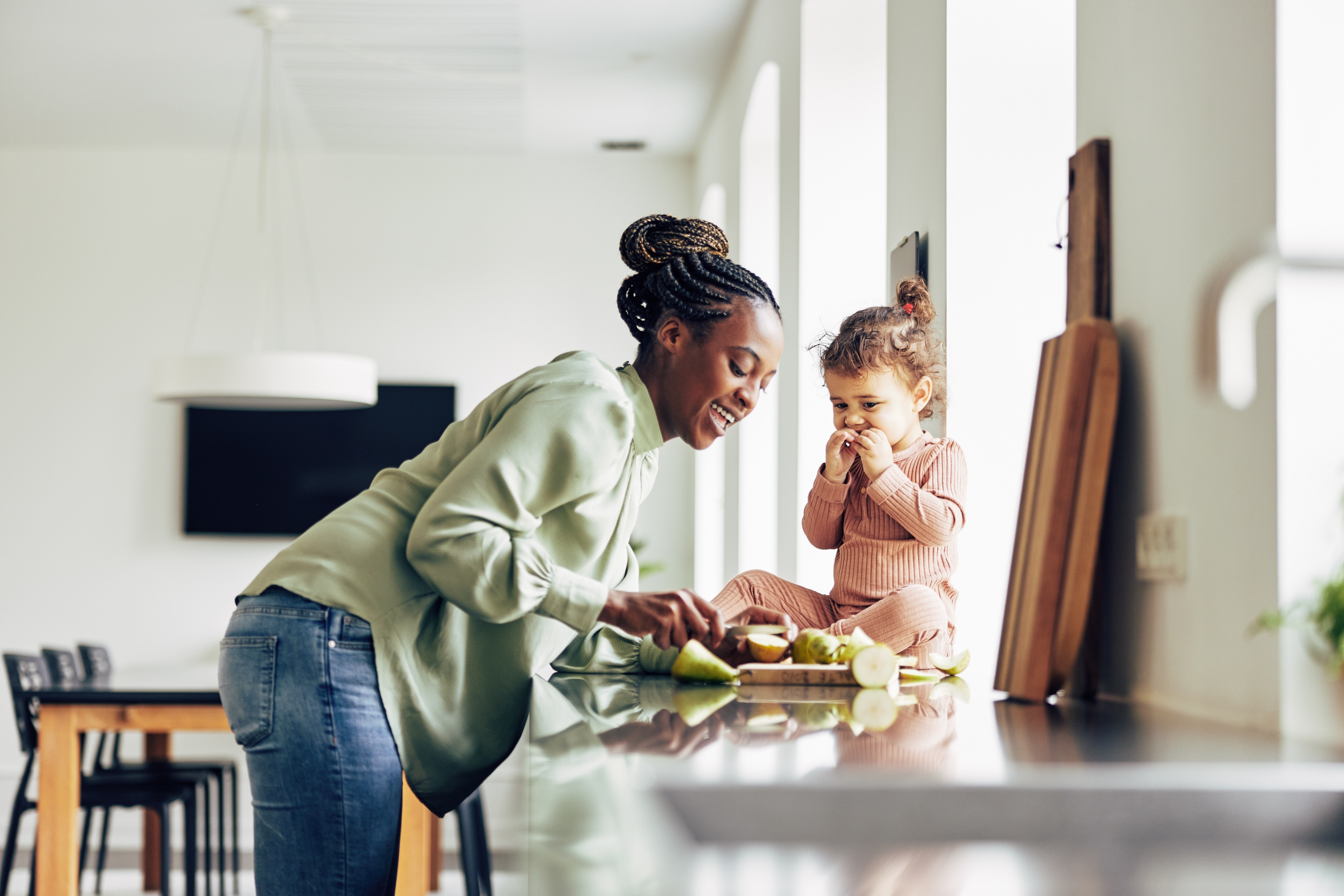 Une mère de race noire coupe des fruits pour sa petite fille dans la cuisine tout en souriant. La petite est assise sur le comptoir et mange les fruits. 