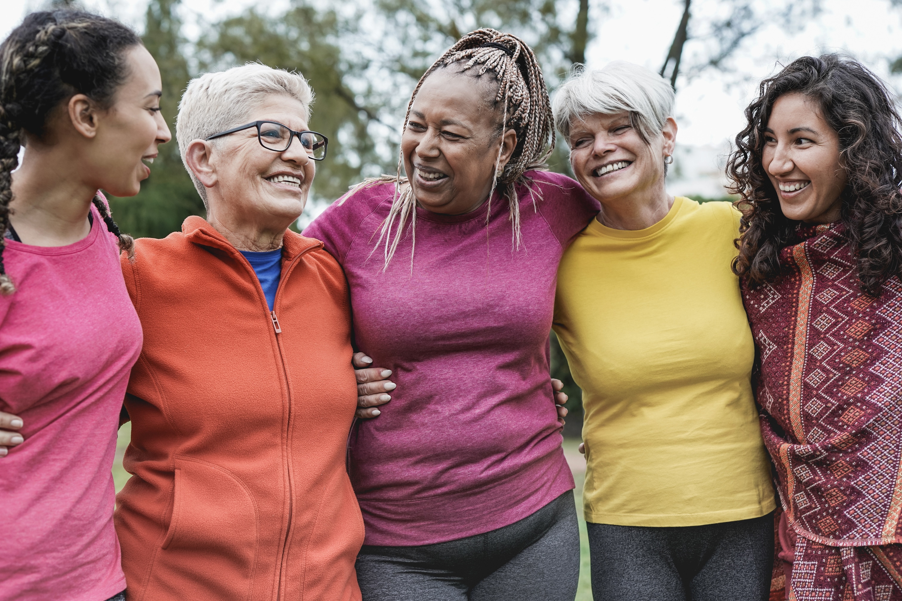 Five women of different backgrounds and different ages have their arms around each other outside. They are all smiling together.