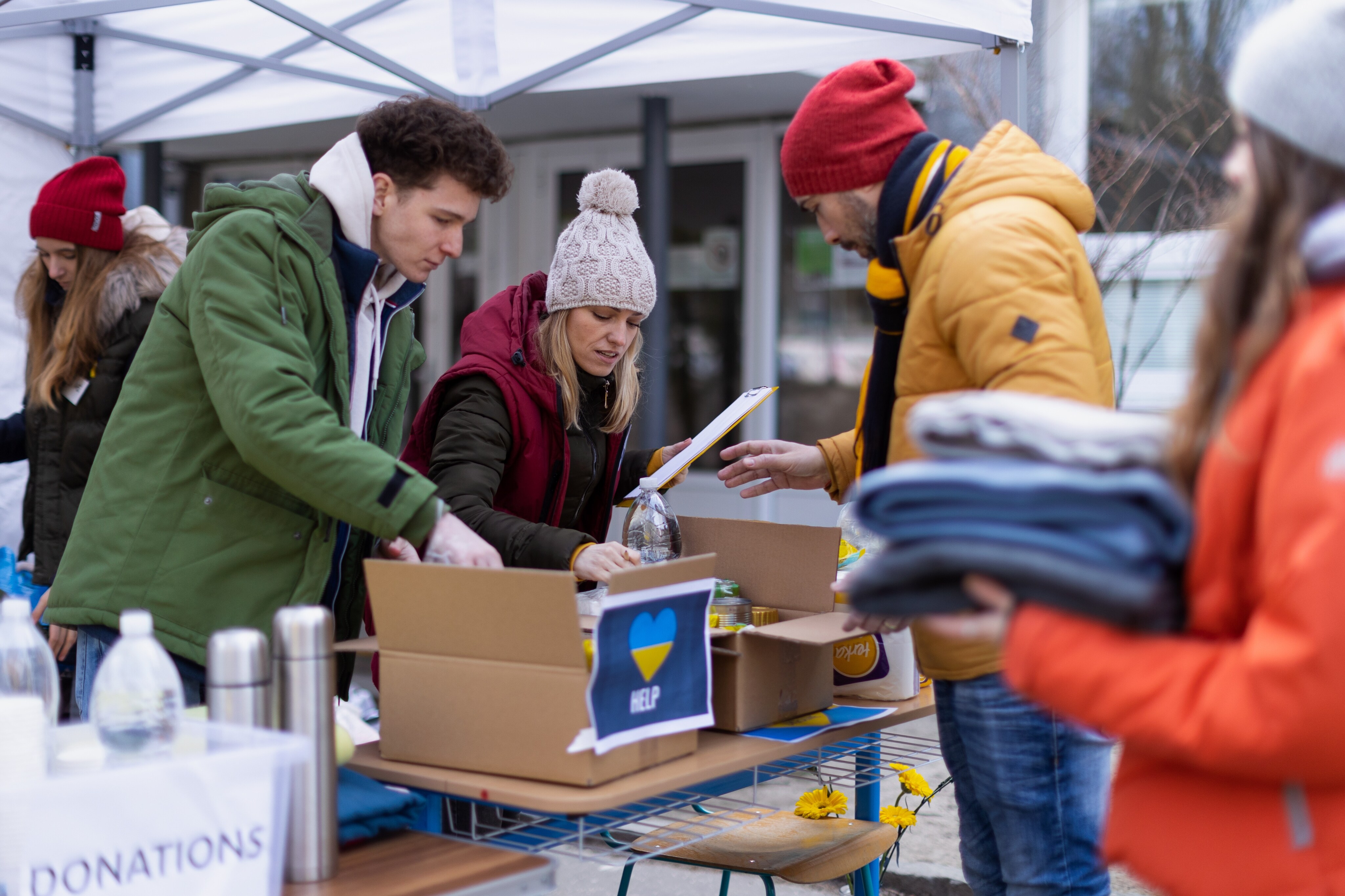 Two men and one woman are looking at merchandise inside boxes. They are at an outdoor fundraiser event for Ukraine. There are two other women nearby.