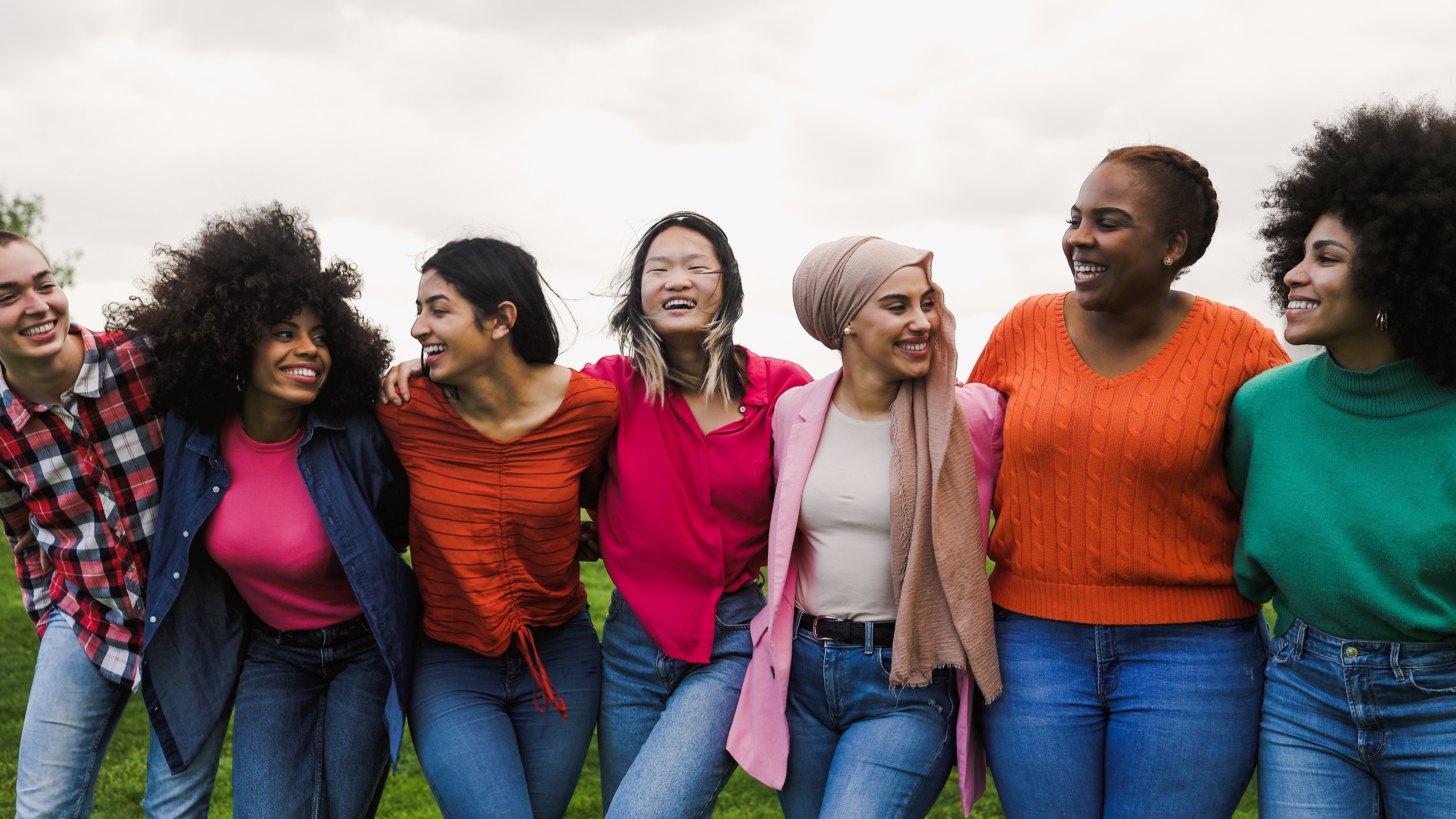 Seven women of different backgrounds have their arms around each other outside. They are dressed in bright colours and smiling.