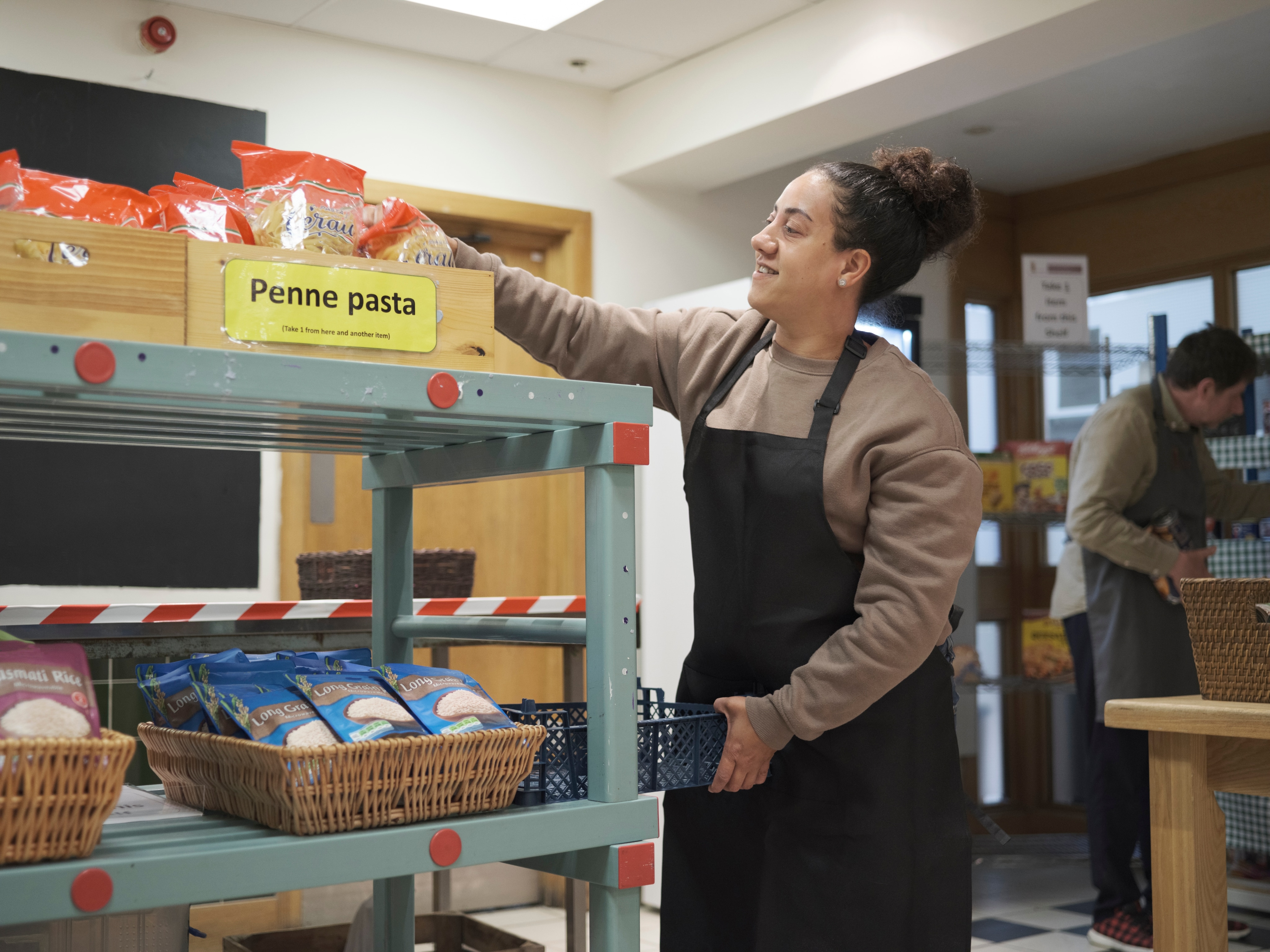 A female food bank employee is grabbing pasta from a shelf. There is a male employee grabbing food behind her, too.
