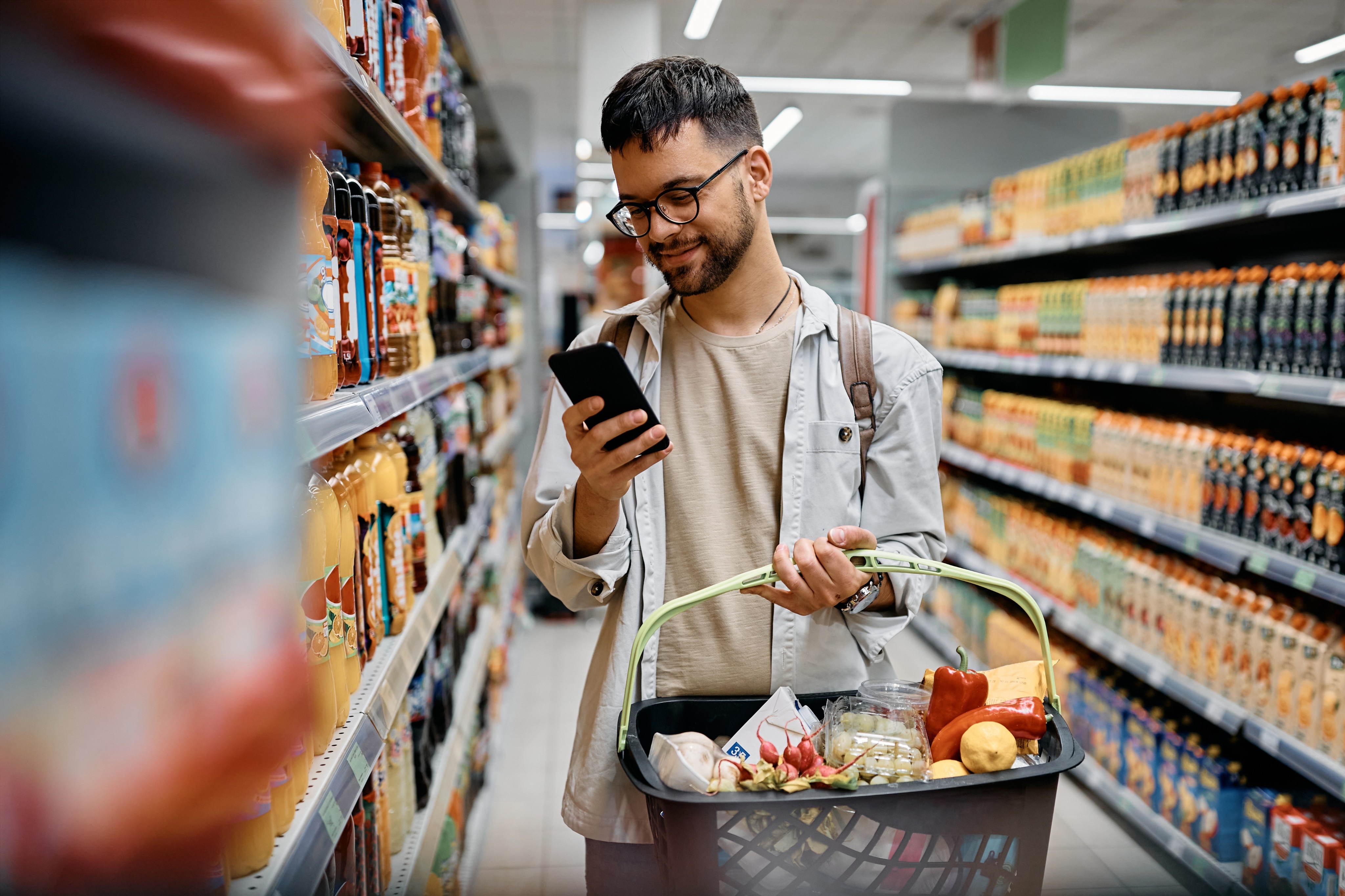 A man is smiling at his phone while holding a basket of groceries. He is standing in a food aisle of a grocery store. 