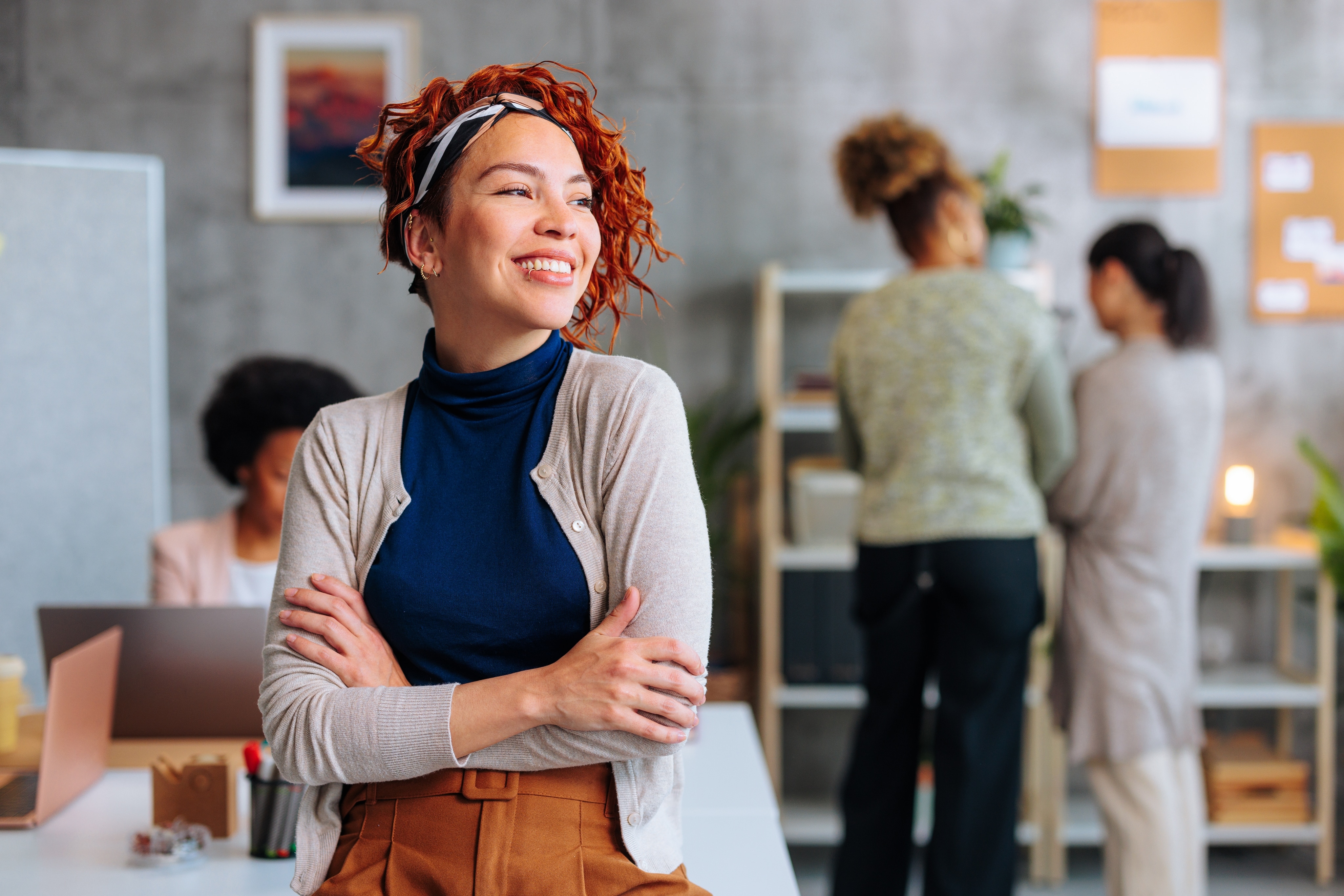 Une femme aux cheveux bouclés orange croise les bras et sourit dans un bureau. Trois autres femmes se trouvent derrière elle. 