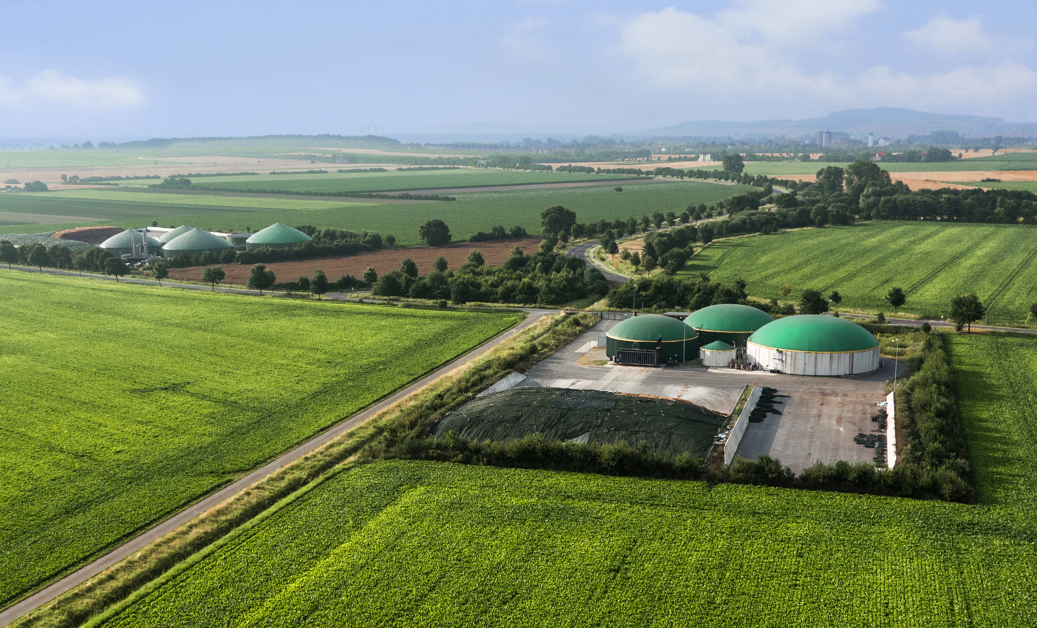 A few factory buildings are surrounded by paved roads, green grass, green trees, and blue sky. 