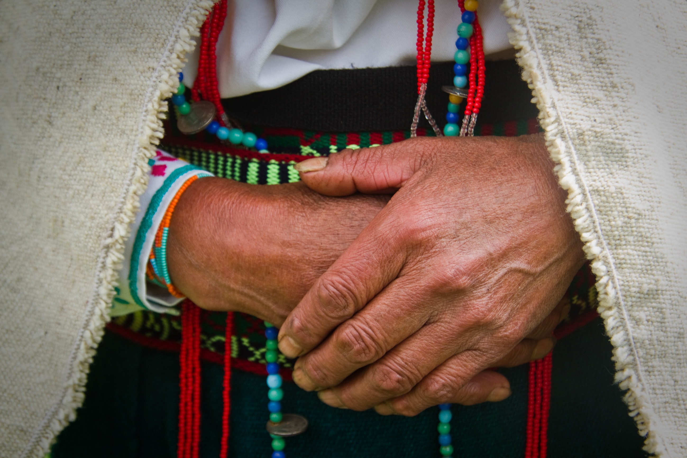 An Indigenous person holds their hands together while wearing cultural clothing. This includes colourful beads, a colourful belt, and a woven shawl. 