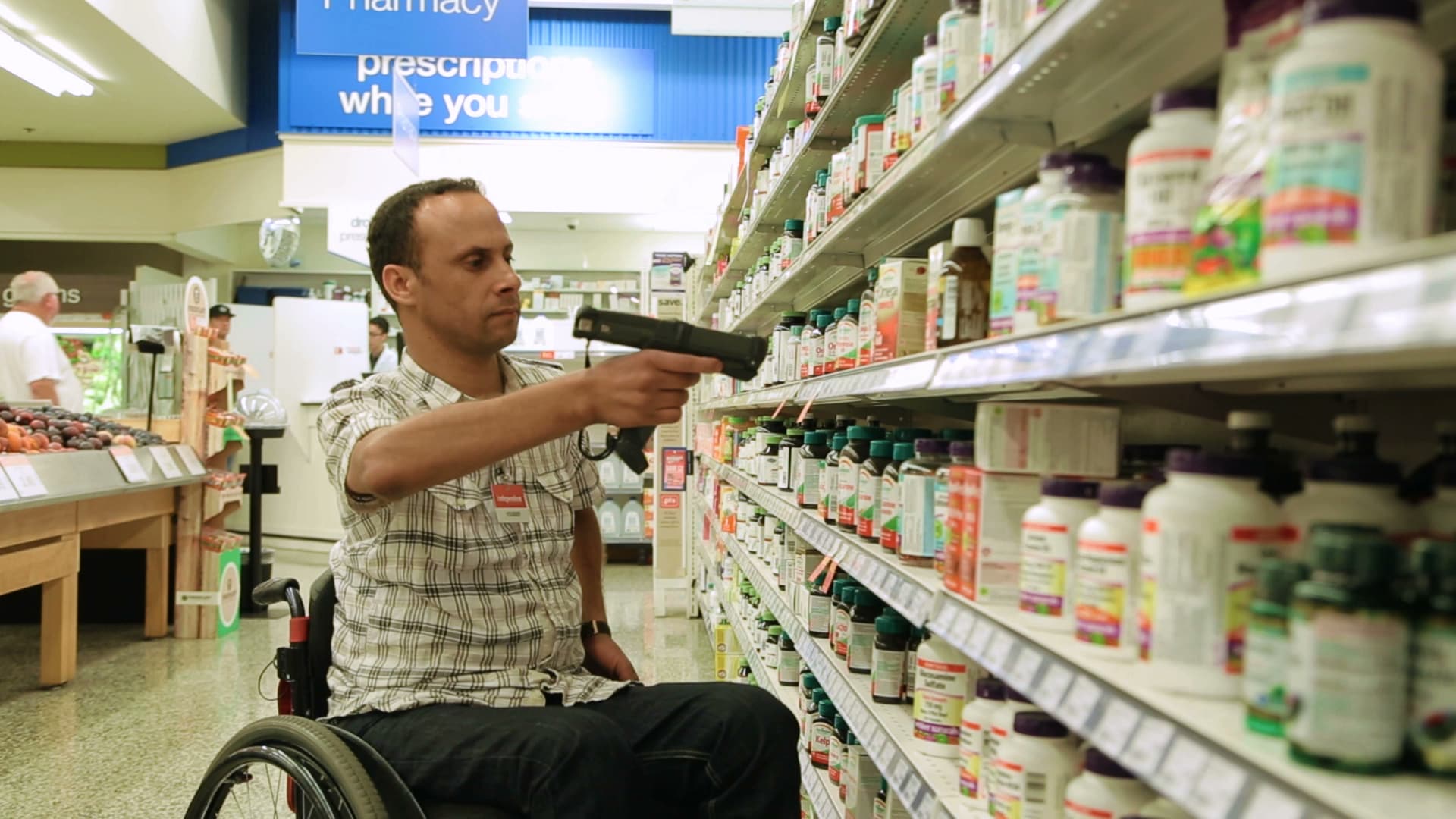 A male employee is sitting in a wheelchair. He is scanning vitamins in a pharmacy aisle at a grocery store. 