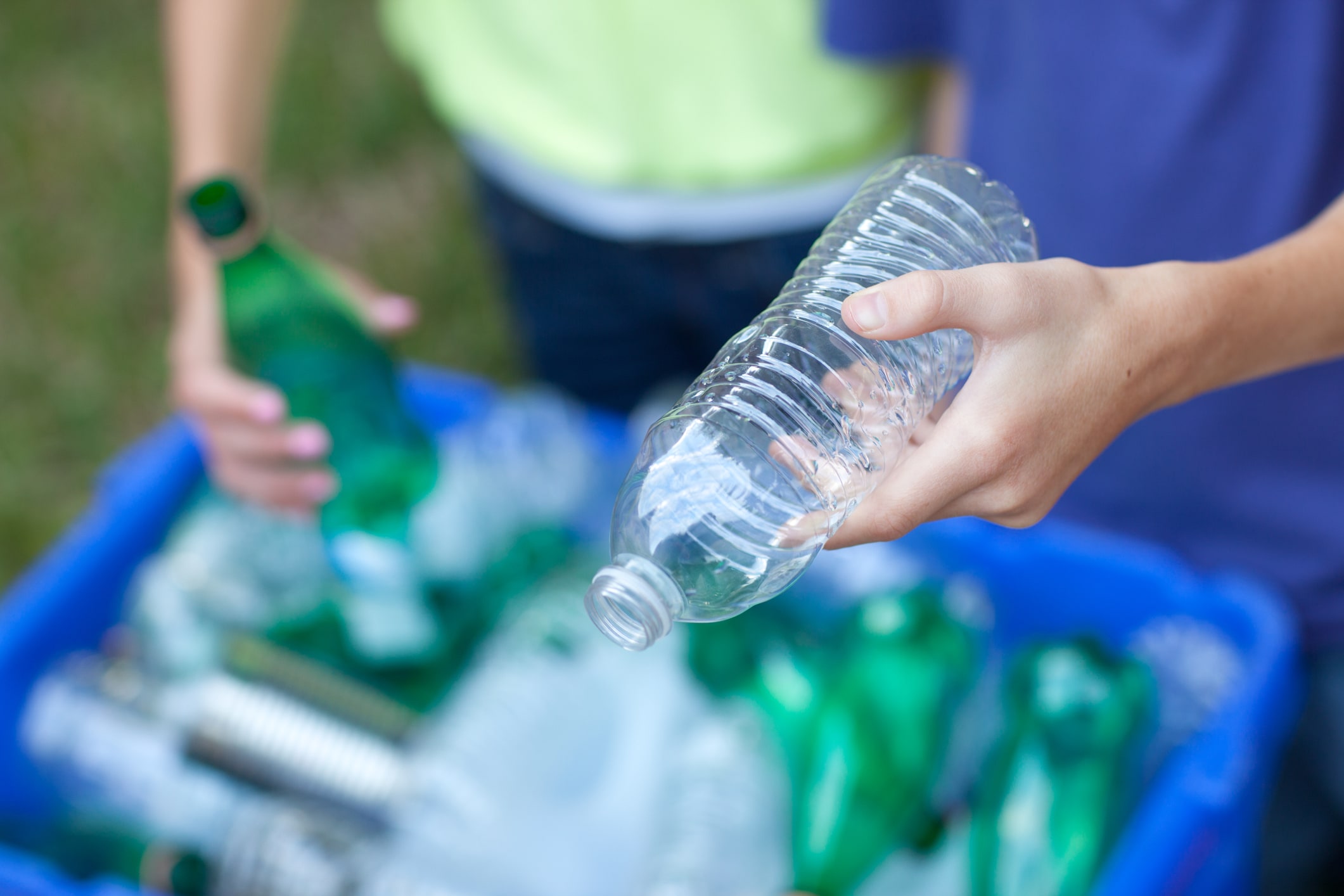 A person holds a green glass bottle while another person holds a clear plastic bottle. They are placing the bottles in a recycling bin.