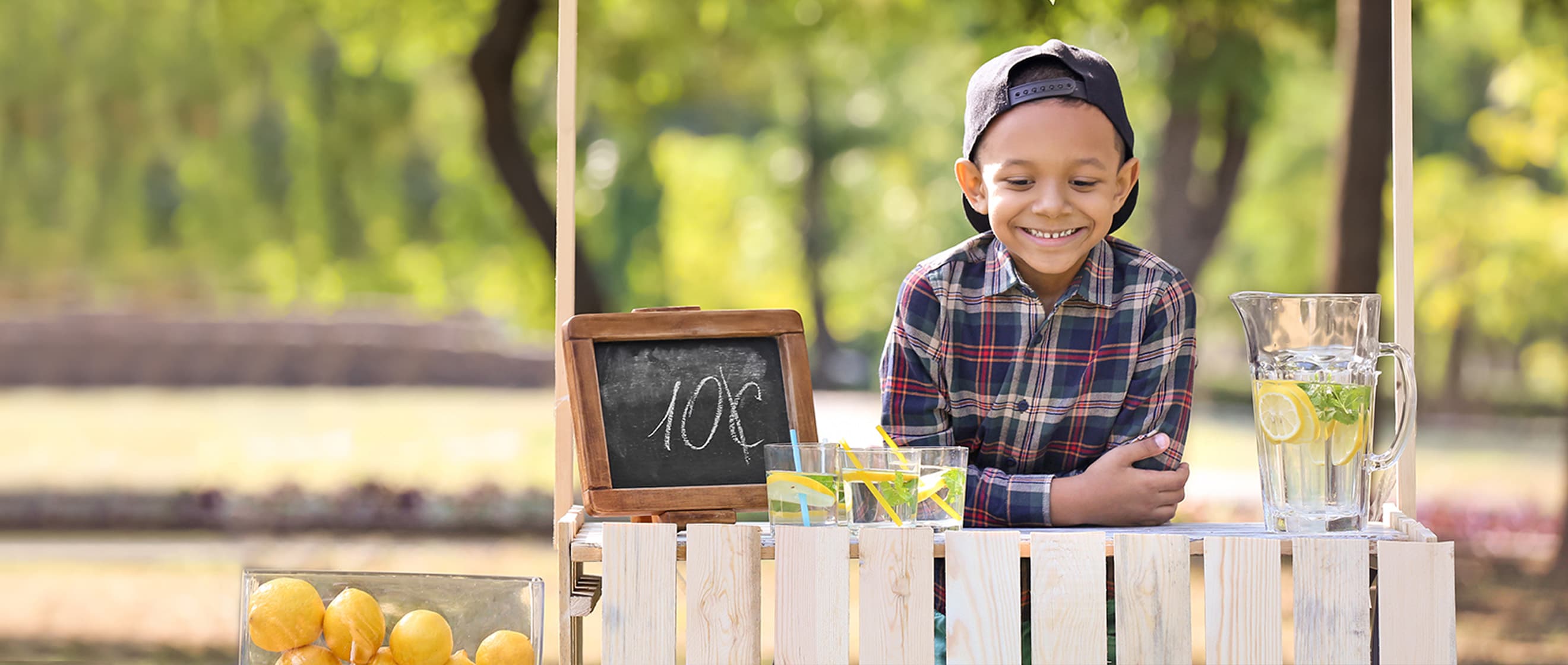 A young boy sits behind a lemonade stand placed outside in a park with trees.