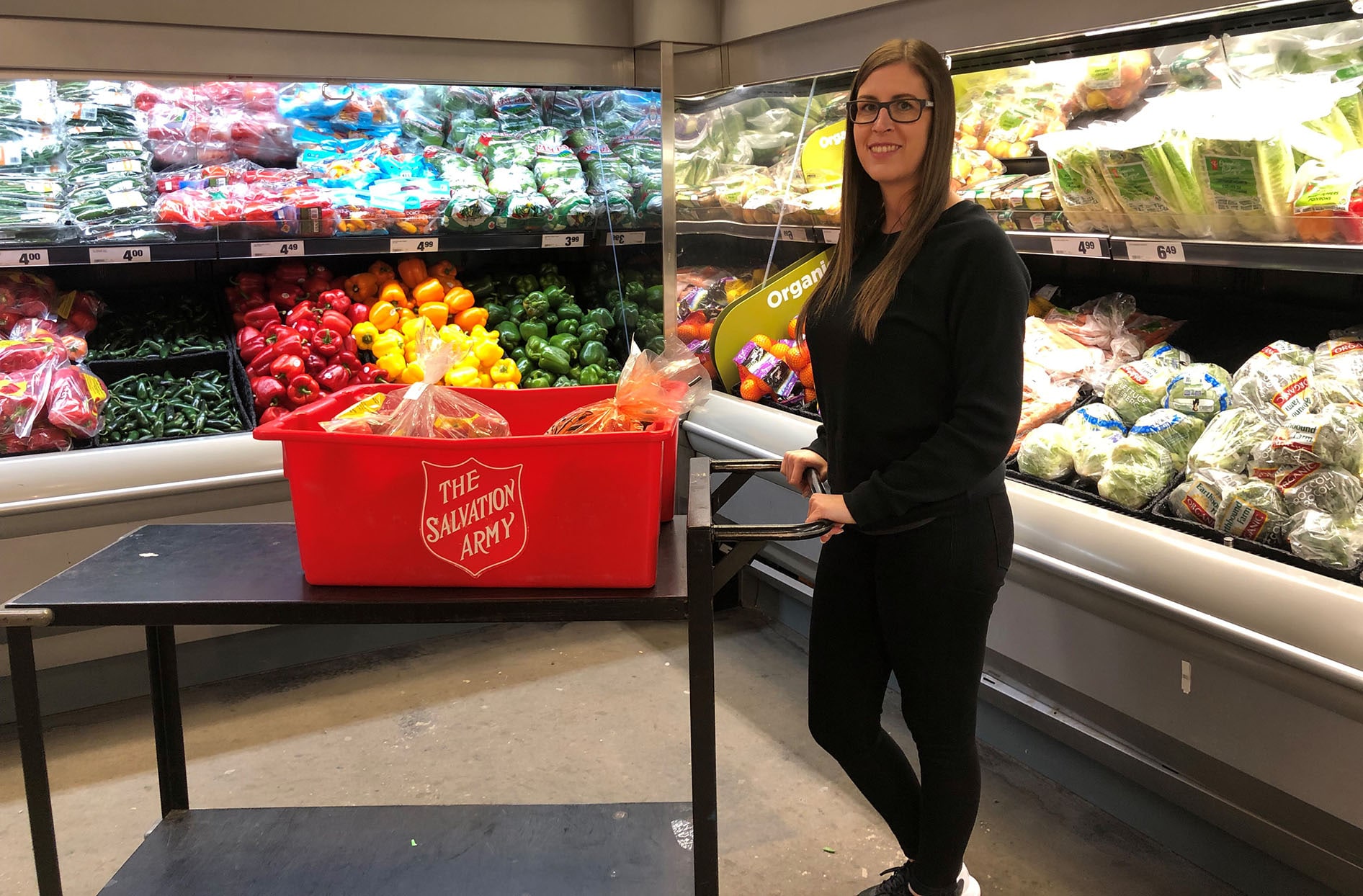 Amanda stands inside of her store pushing a Salvation Army basket with food inside it. 