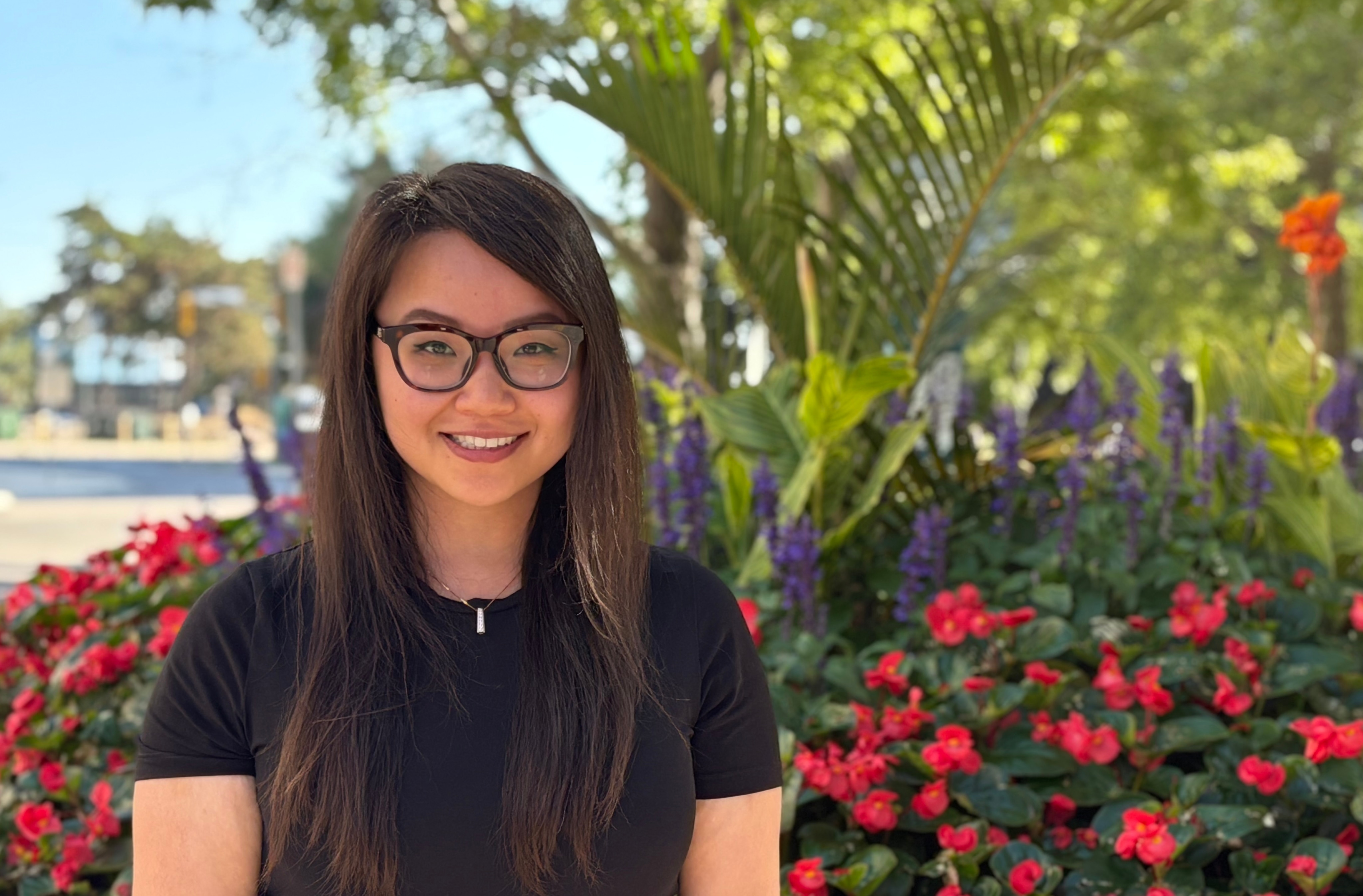 A photo of Christina standing outside with flowers and trees behind her.  
