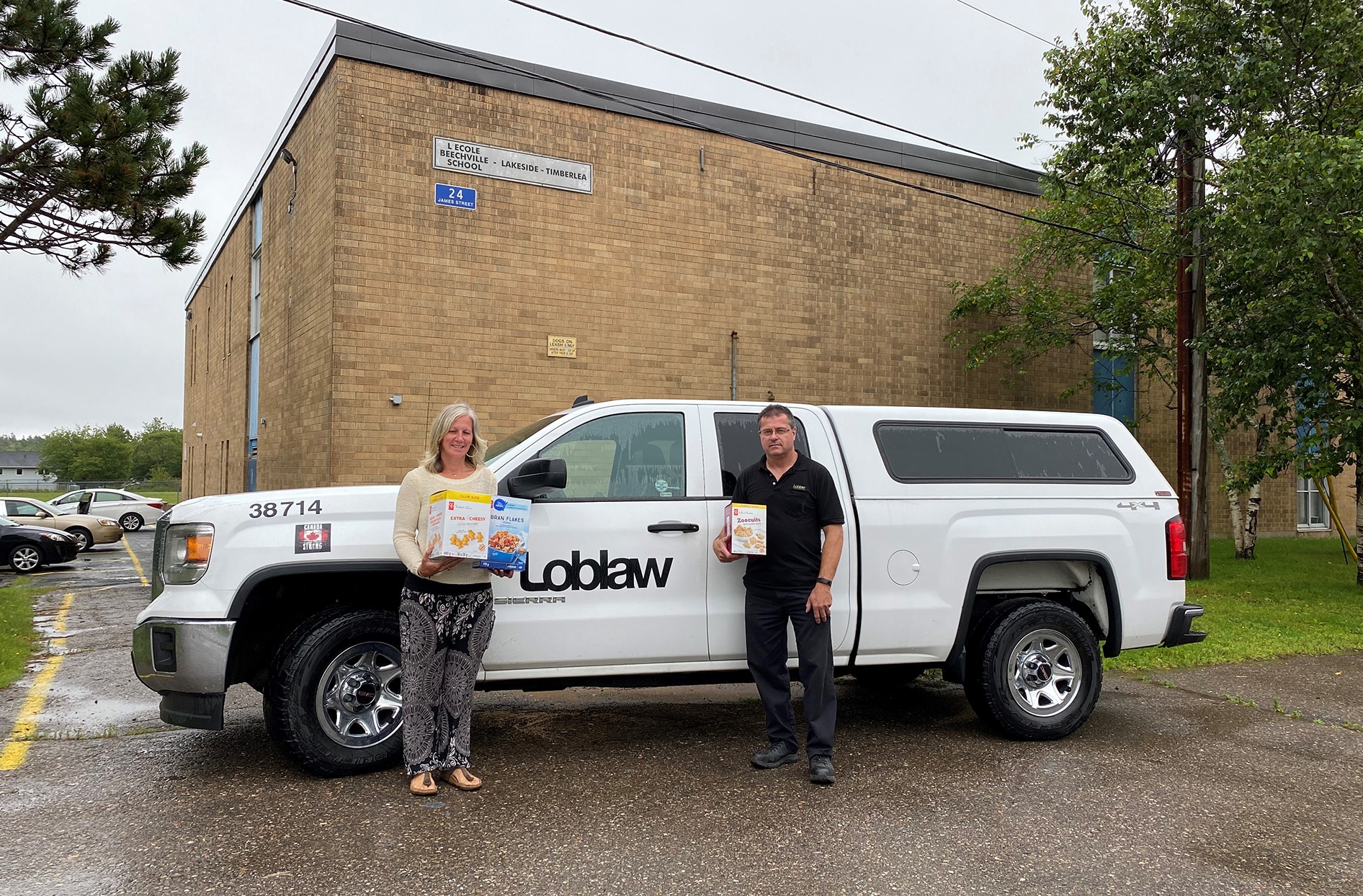 David se tient à côté d’un camion Loblaw avec la vice-directrice de l’école, et les deux tiennent des produits PC. 