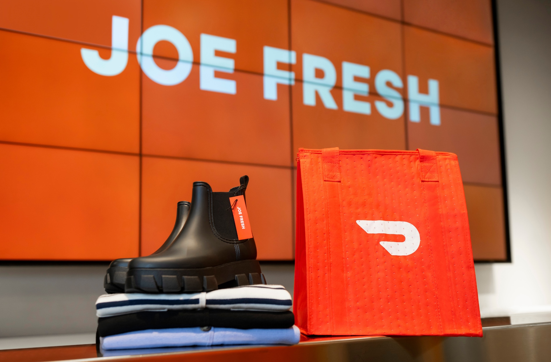 Black ankle boots displayed on folded clothing beside a red DoorDash delivery bag, with a large “JOE FRESH” sign on an orange screen in the background.