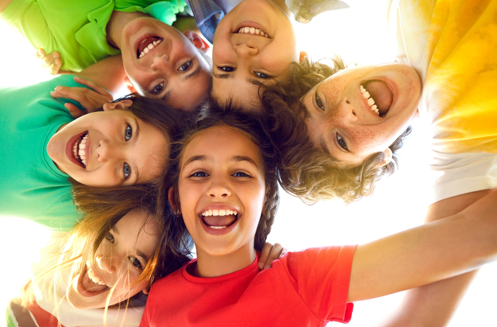 Six children smiling and laughing together in a circle outdoors, viewed from below against a bright sunny sky.
