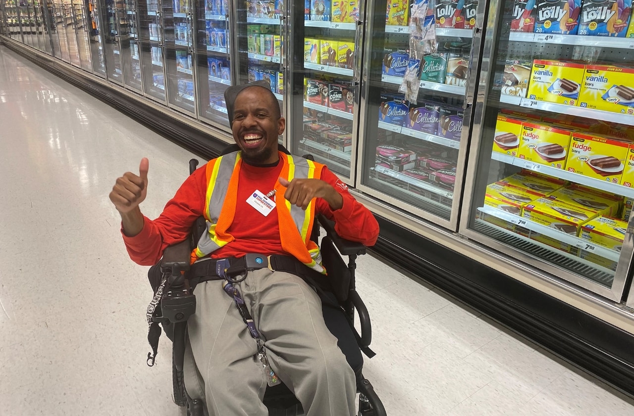 Un homme souffrant de paralysie cérébrale est  assis dans son fauteuil roulant dans une allée d’épicerie en souriant devant la caméra