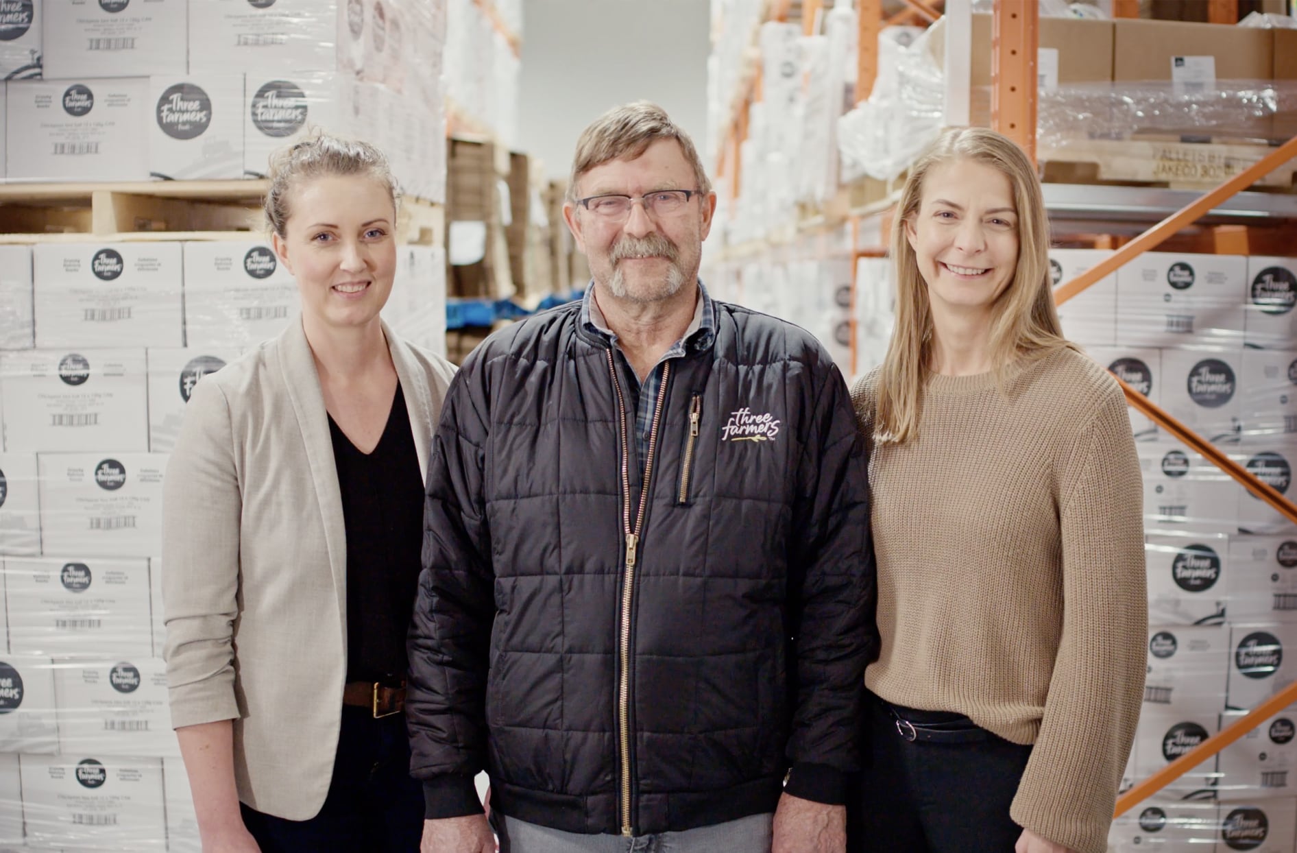 Founders of Three Farmers Foods Natasha and Elysia Vandenhurk posing proudly with their father in front of their packaged products inside a warehouse. 