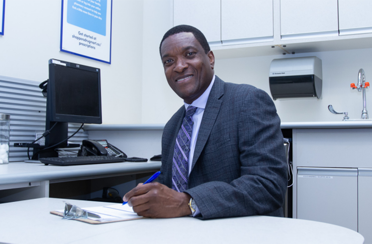 Smiling man sitting at desk, pen in left hand, paper and clipboard on desk