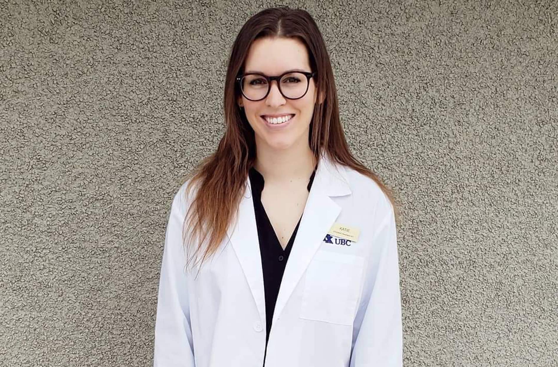 Katie stands in front of a wall wearing her pharmacy lab coat 