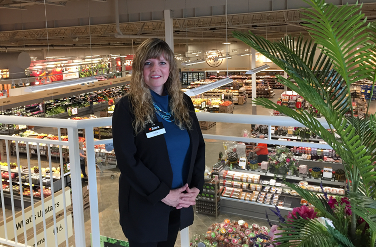 Lynda stands on the second level of her store with her hands together, smiling into the camera