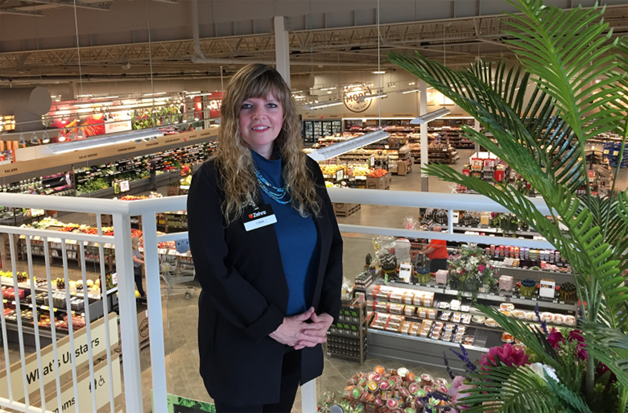 Lynda se tient au deuxième étage de son magasin, les mains jointes, souriant à la caméra.