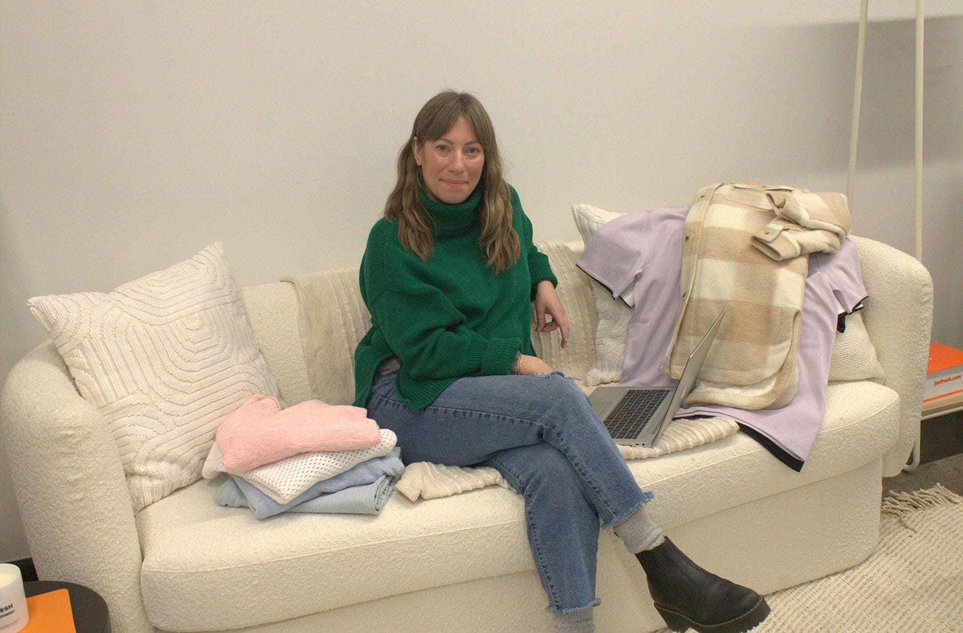 Melissa sits on a couch inside the Joe Fresh office with her laptop on her lap and clothing samples next to her. 