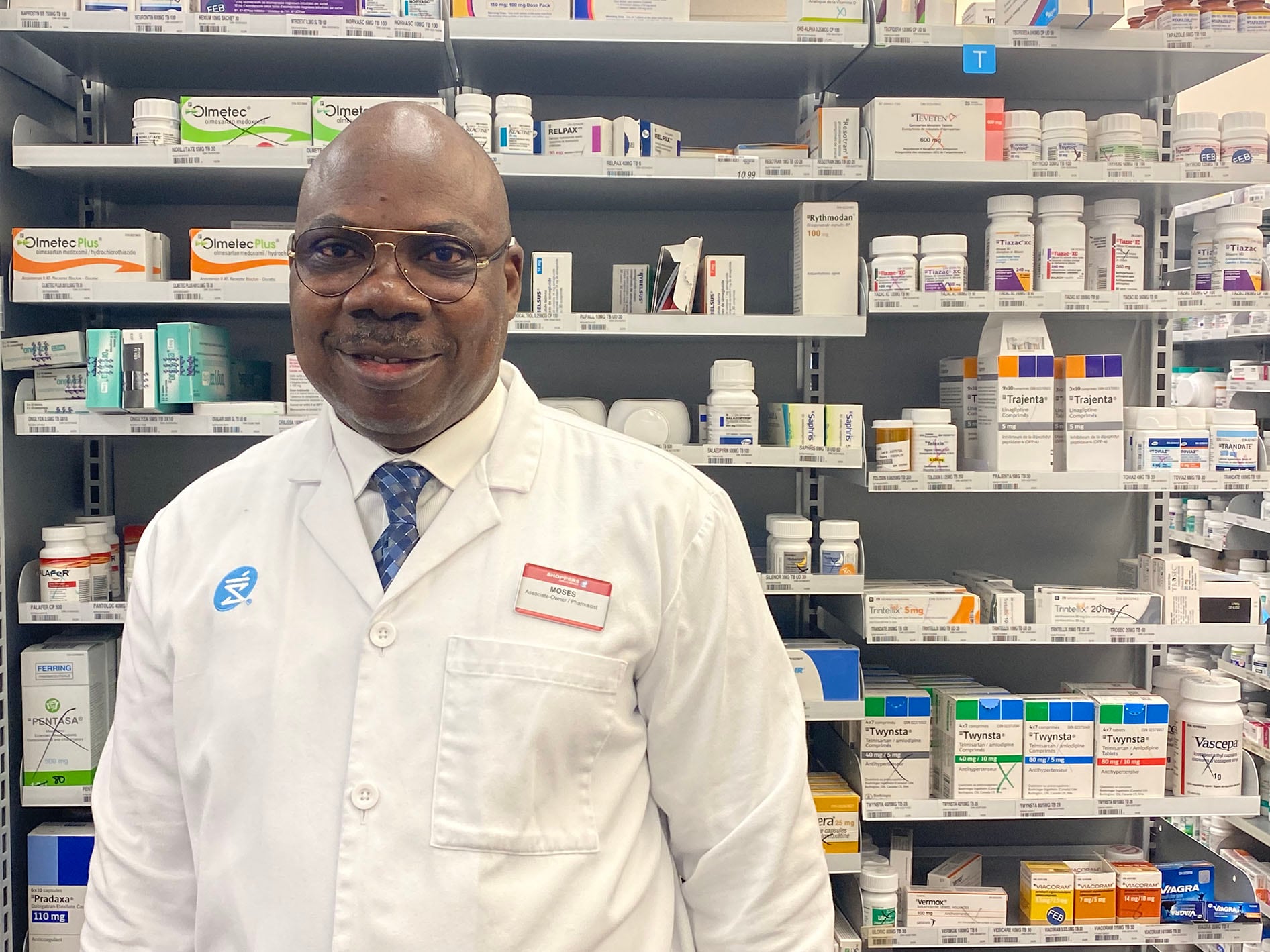 Moses stands wearing his lab coat behind the pharmacy counter.   