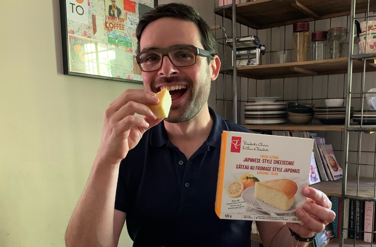 Man sitting at dining table about to bite into a piece of cheesecake in one hand, holding box of cheesecake in other hand