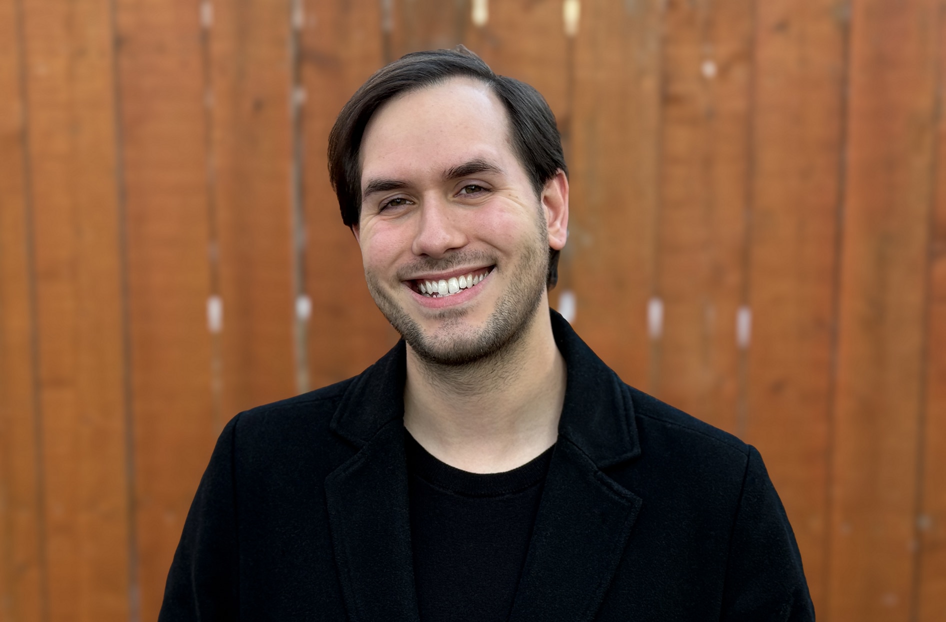 Nicolas is standing in front of a brown fence smiling. 