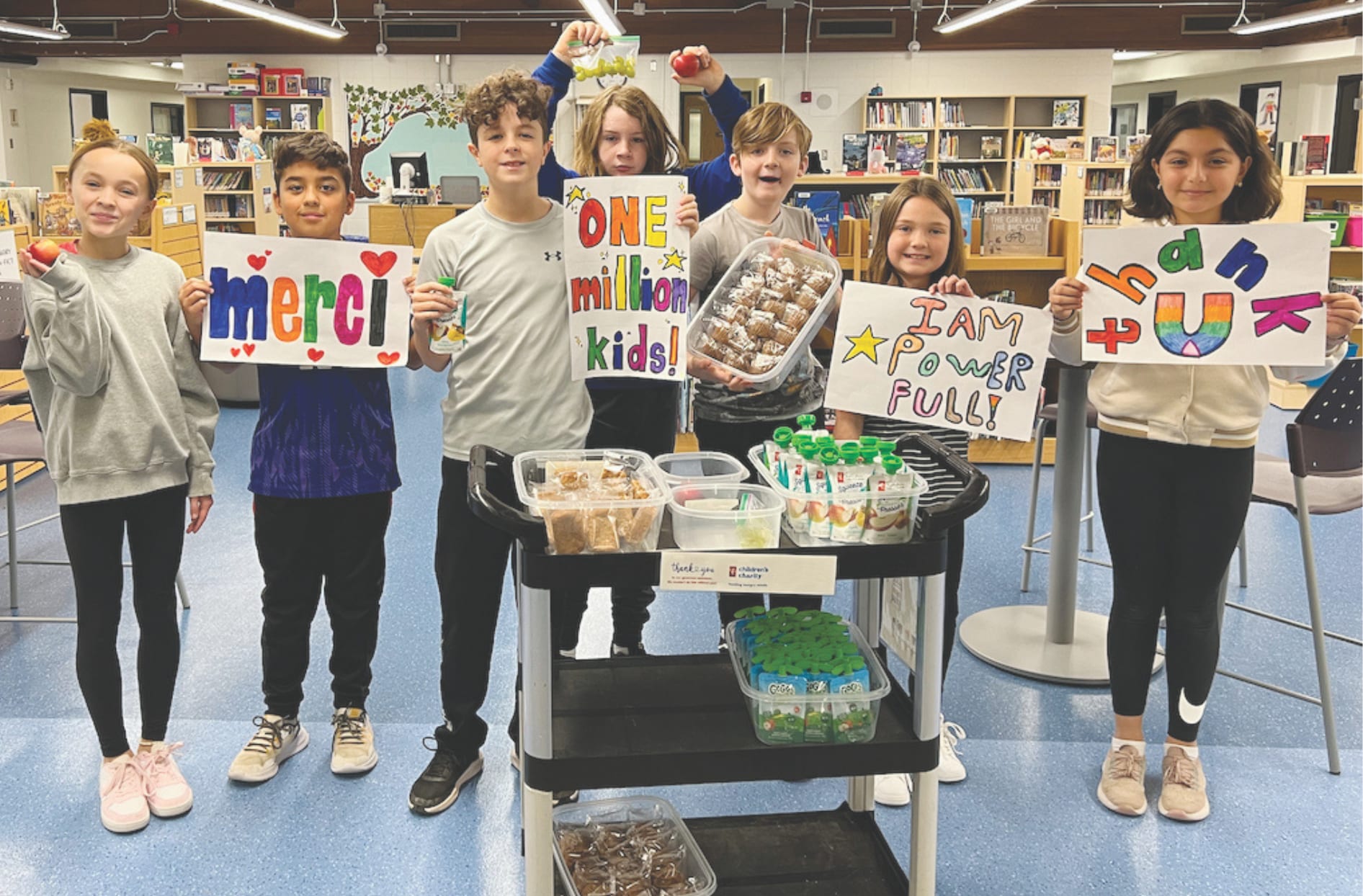 Un groupe d’enfants qui se tiennent à l’intérieur d’une école et qui tiennent des affiches pour remercier la « Fondation » avec un chariot d’aliments devant eux. 