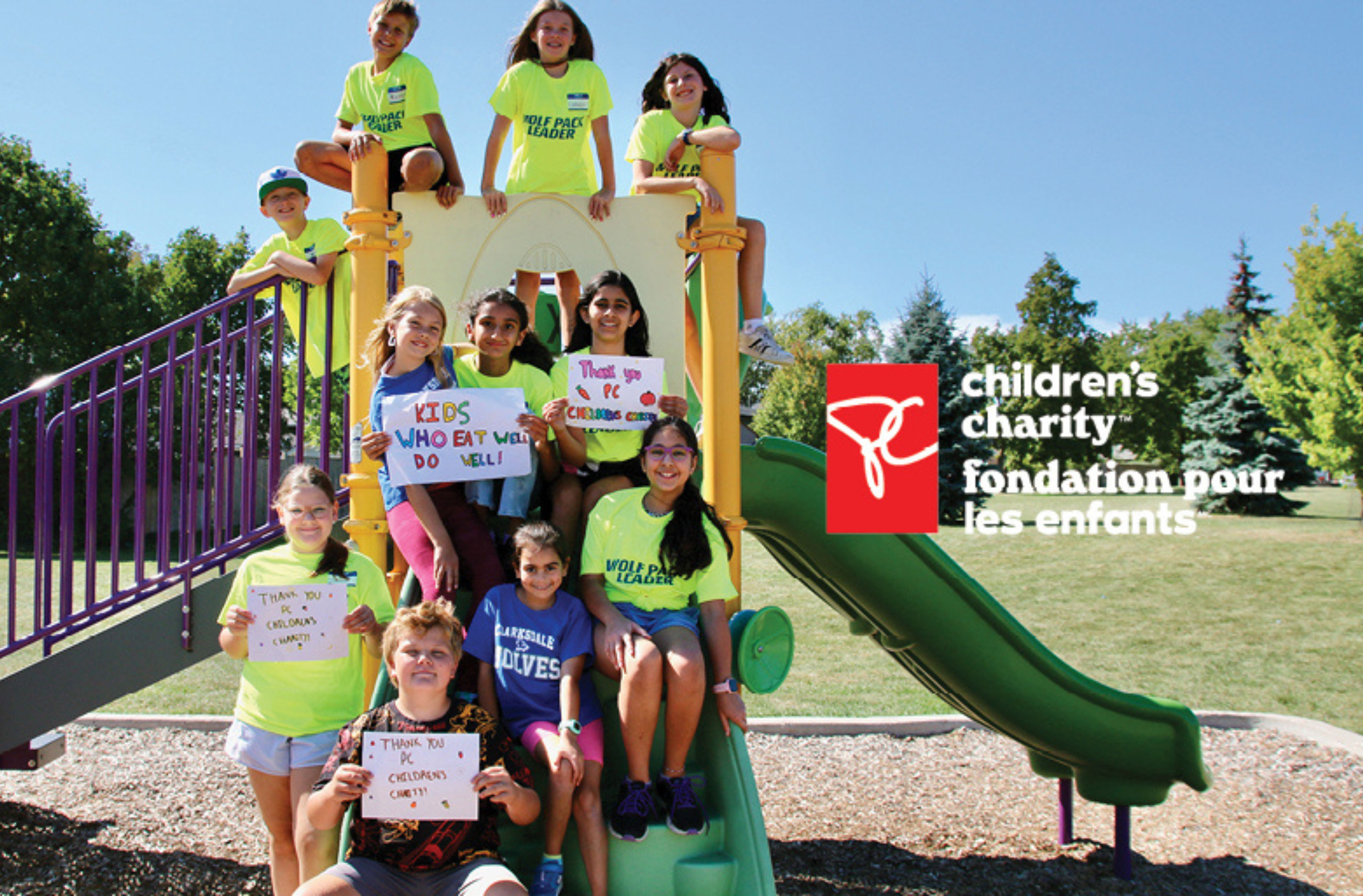 Smiling children on a playground hold signs thanking PC Children’s Charity.