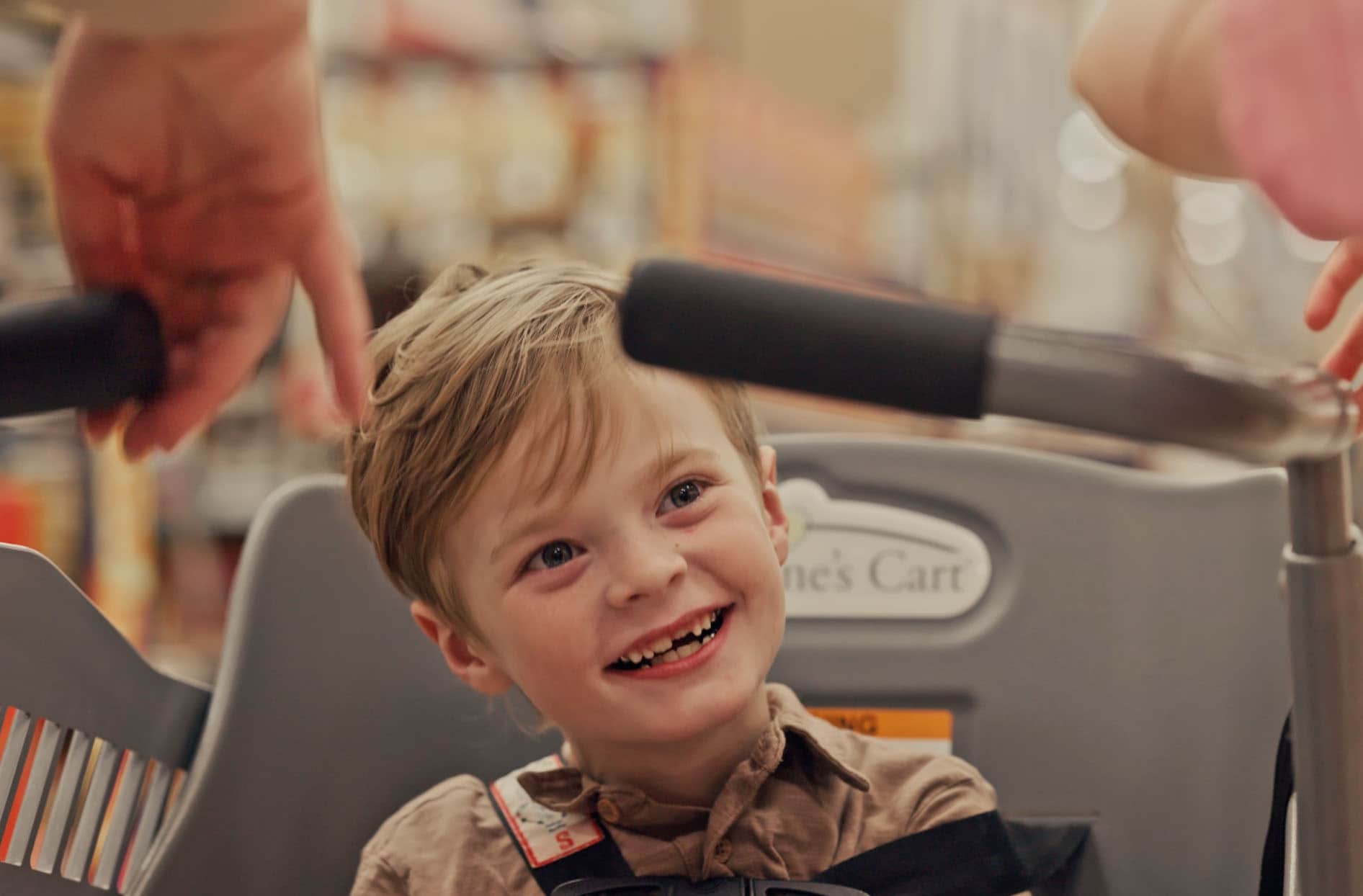 A young boy with mobility issues smiling while shopping with his family using a Caroline’s Cart. 