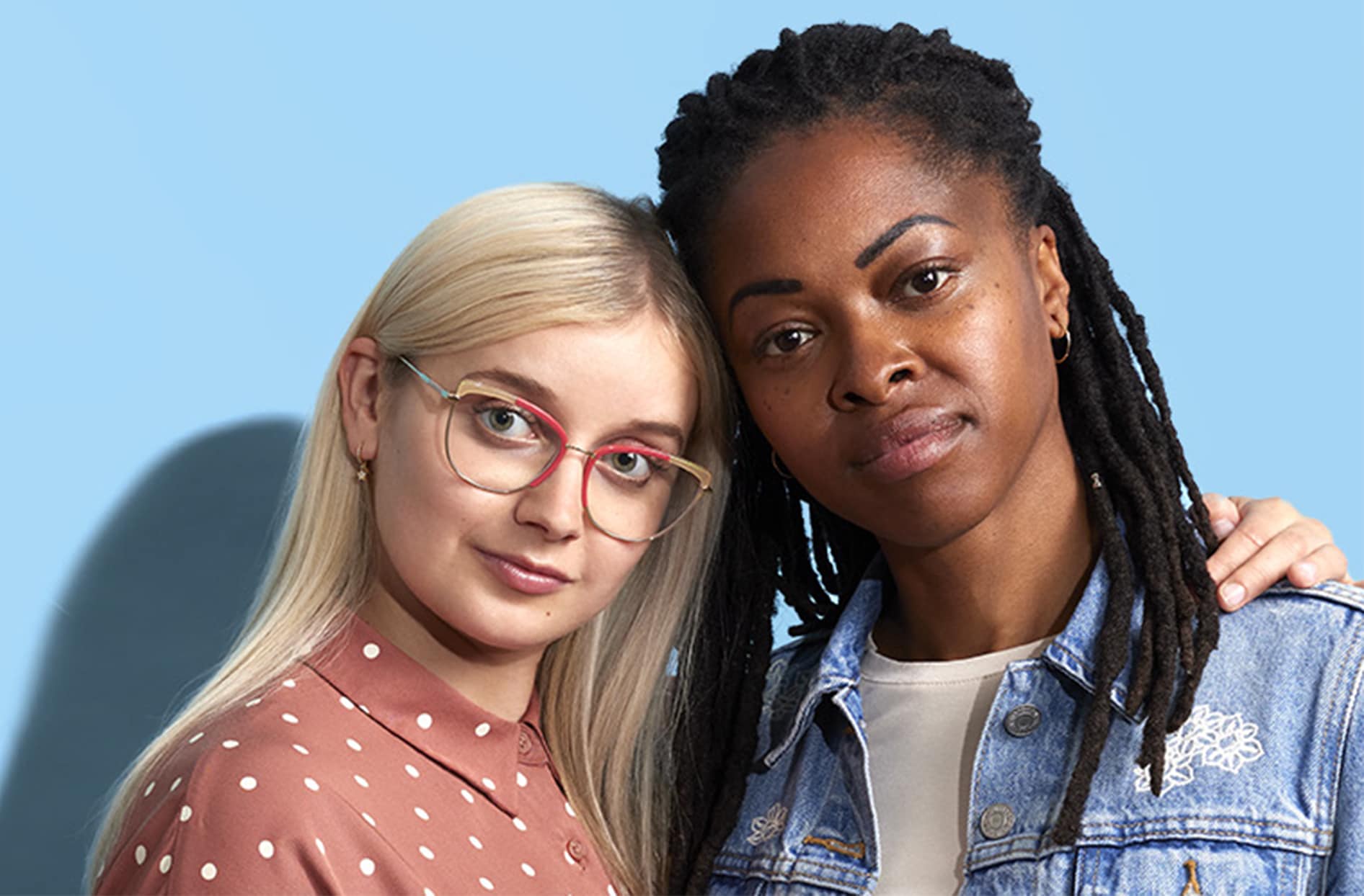 Two women standing close together against a light blue background. One woman has long blonde hair, wears glasses, and a polka-dot blouse, while the other has dark braided hair and wears a denim jacket.