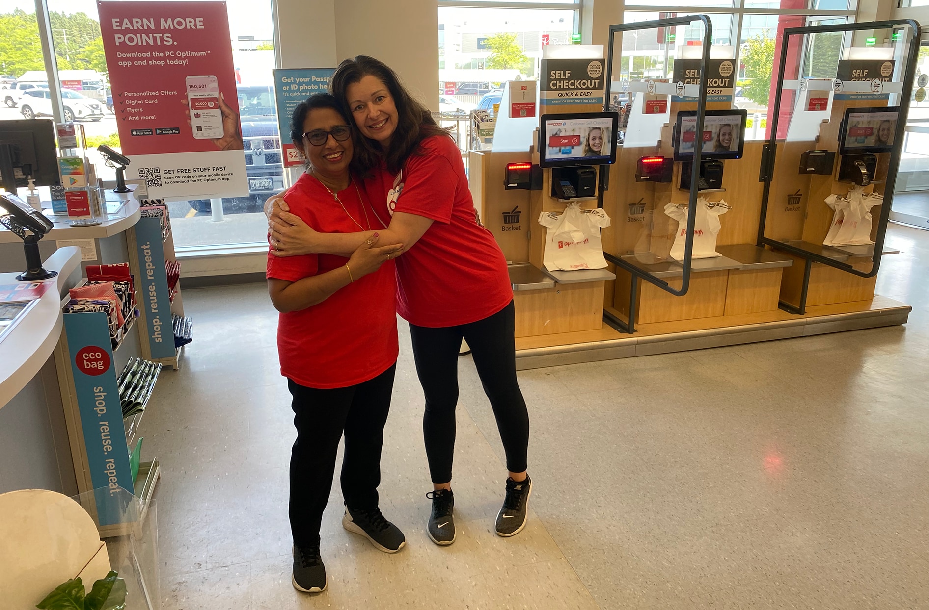 Tina et Linda se prennent dans leurs bras devant les caisses libre-service à l’intérieur de leur magasin Shoppers Drug Mart.