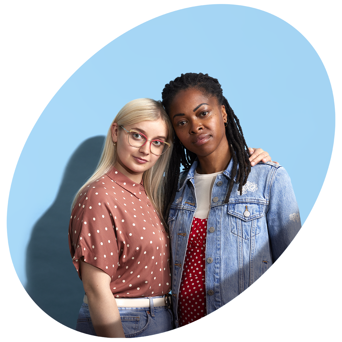 A young White woman, wearing a pink blouse and red glasses, puts her arm around a young Black woman, wearing a denim jacket. They are both standing proud.