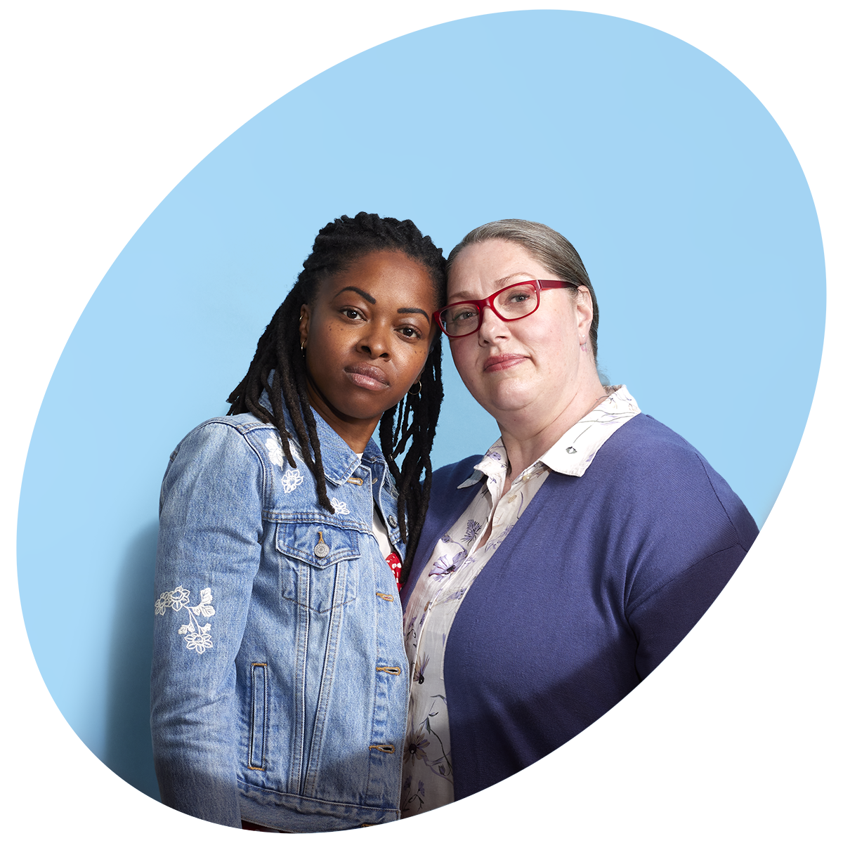 Une jeune femme noire, portant une veste en denim avec des cheveux tressés, se tient à côté d’une femme blanche d’âge moyen, portant un chemisier blanc, un cardigan bleu et des lunettes rouges.