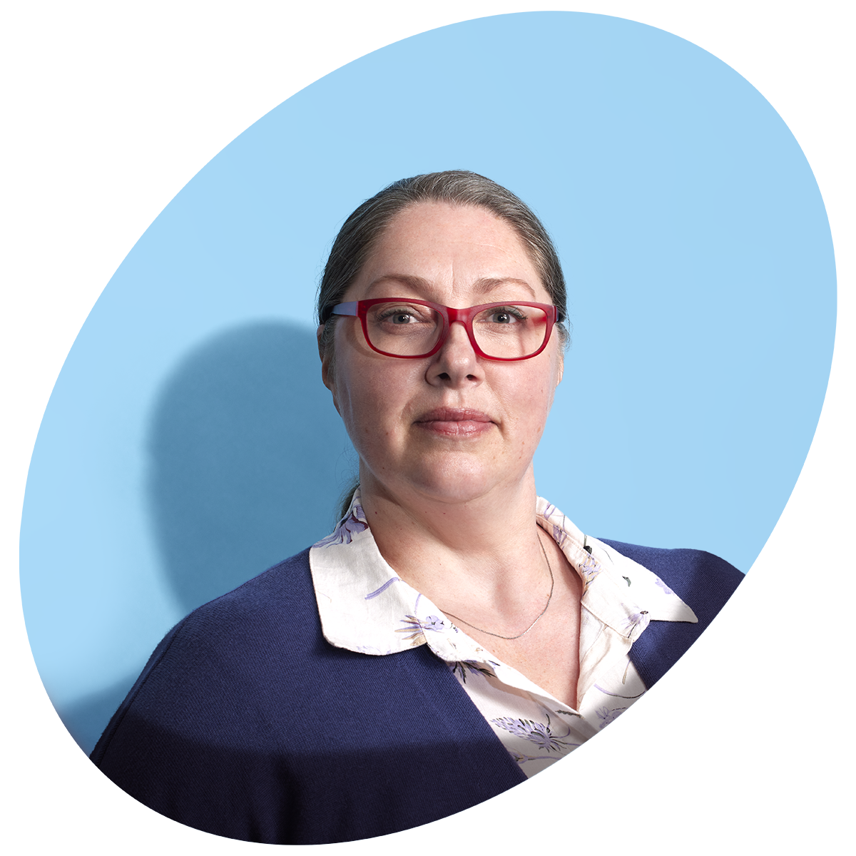 A middle-aged White woman is standing proud. She’s wearing a white blouse, blue cardigan, red glasses, and her hair is tied back.